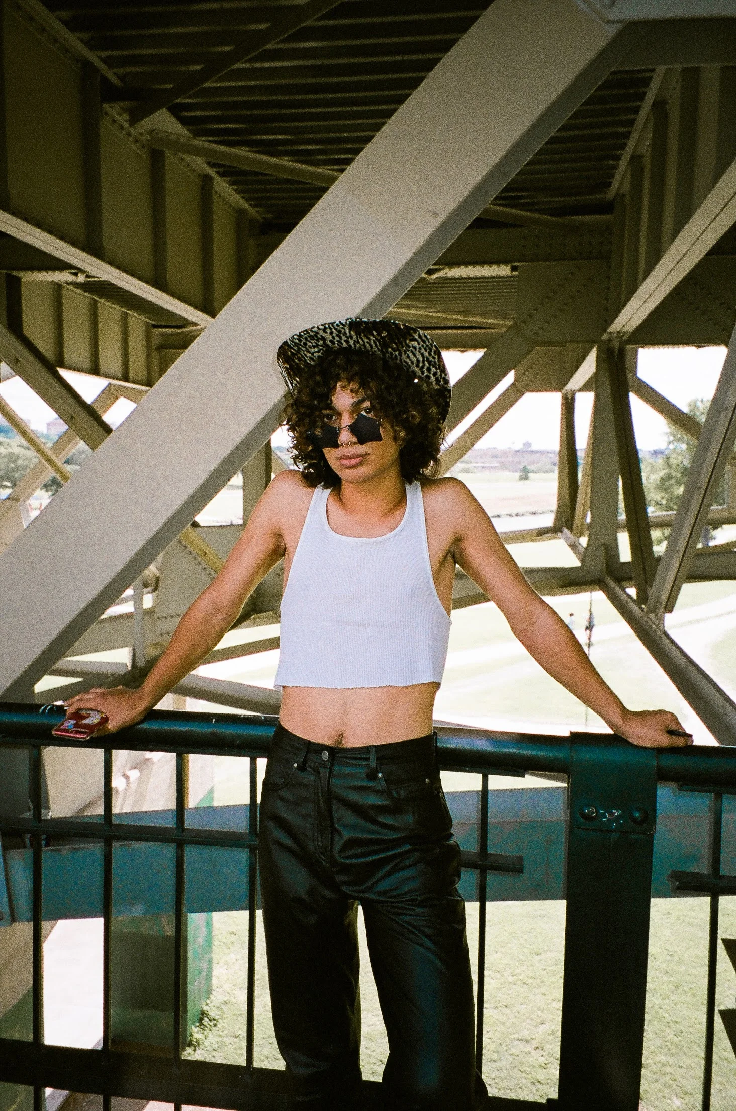 A woman with curly hair wearing a leopard print hat, black sunglasses, a white crop top, and black shiny pants leaning on a railing under a large industrial structure with a grassy field in the background.