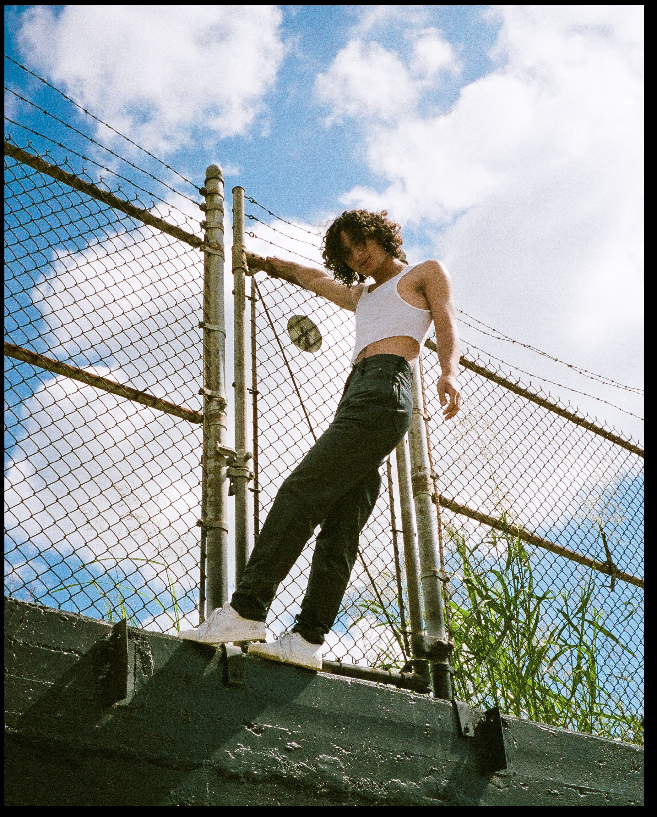 Young woman with curly hair leaning on a chain-link fence, wearing a white crop top, black pants, and white sneakers, standing outdoors under a partly cloudy sky.