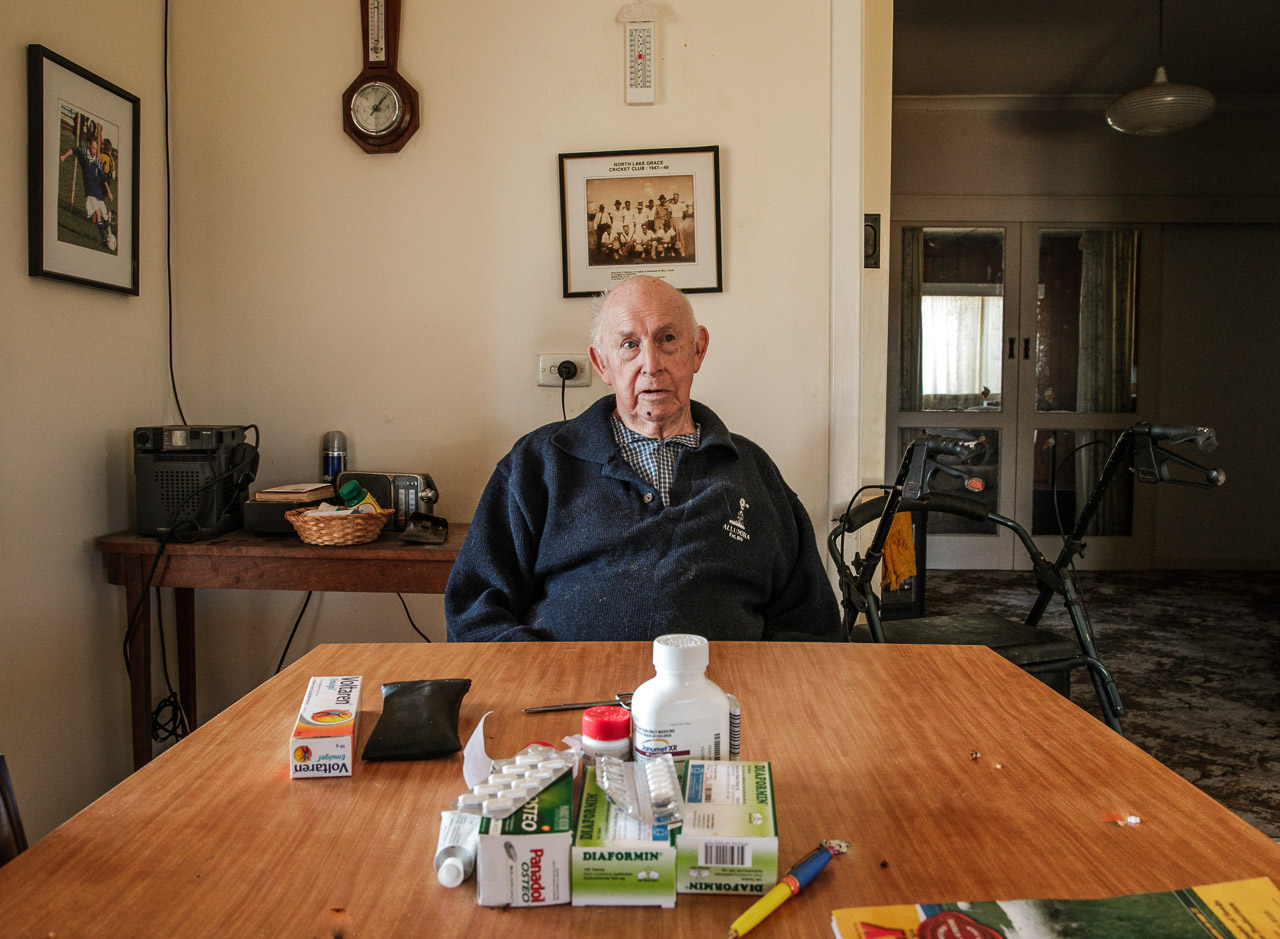 Elderly man at his dining table with his medication, barometer and old cricket team framed photo