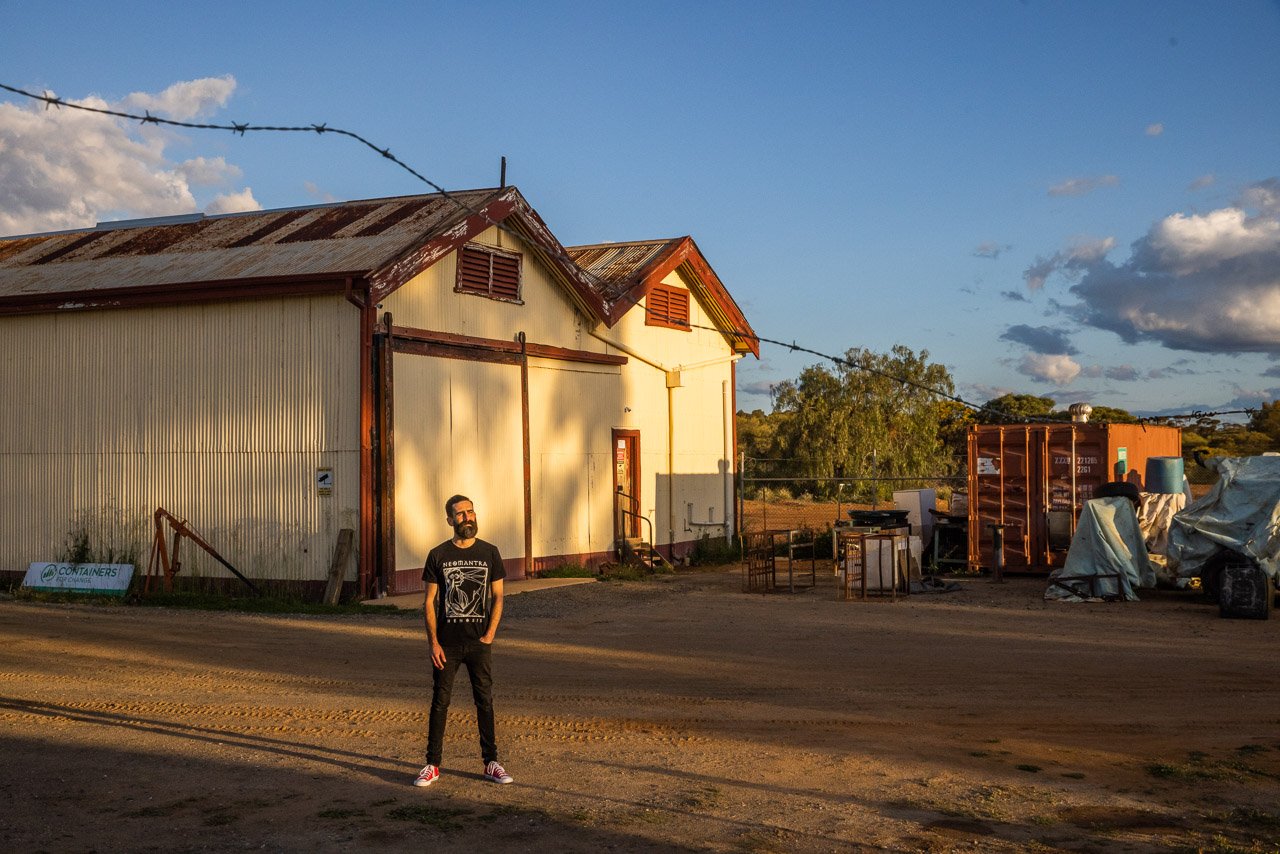 Man wearing black in front of big sheds at sunset