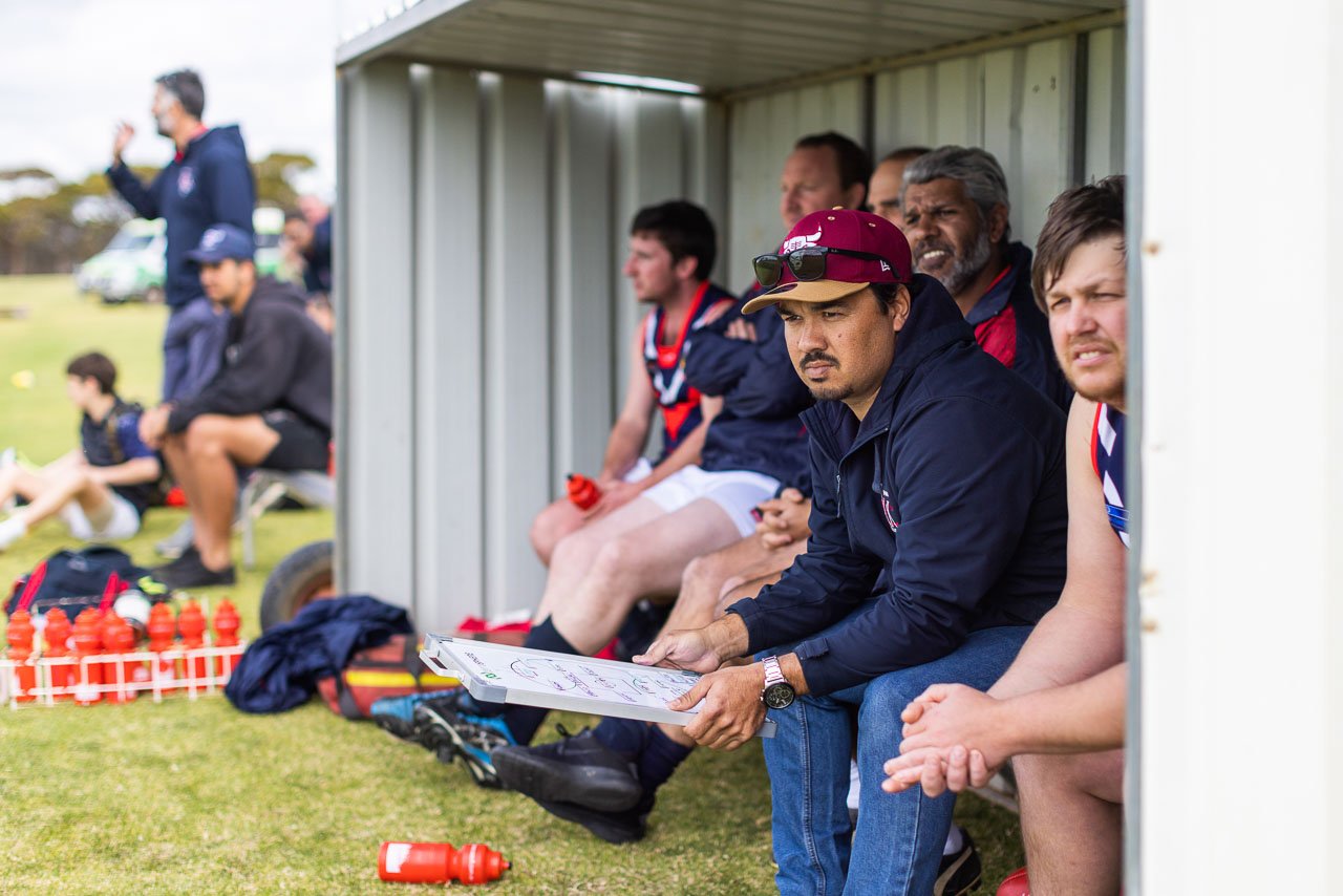 The coach and reserves look on from the sidelines of the footy game in a country town