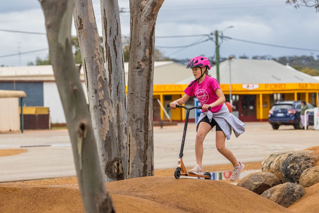 A girl rides her scooter in the public playground in Kulin with the bright yellow Men's Shed and Museum in the background