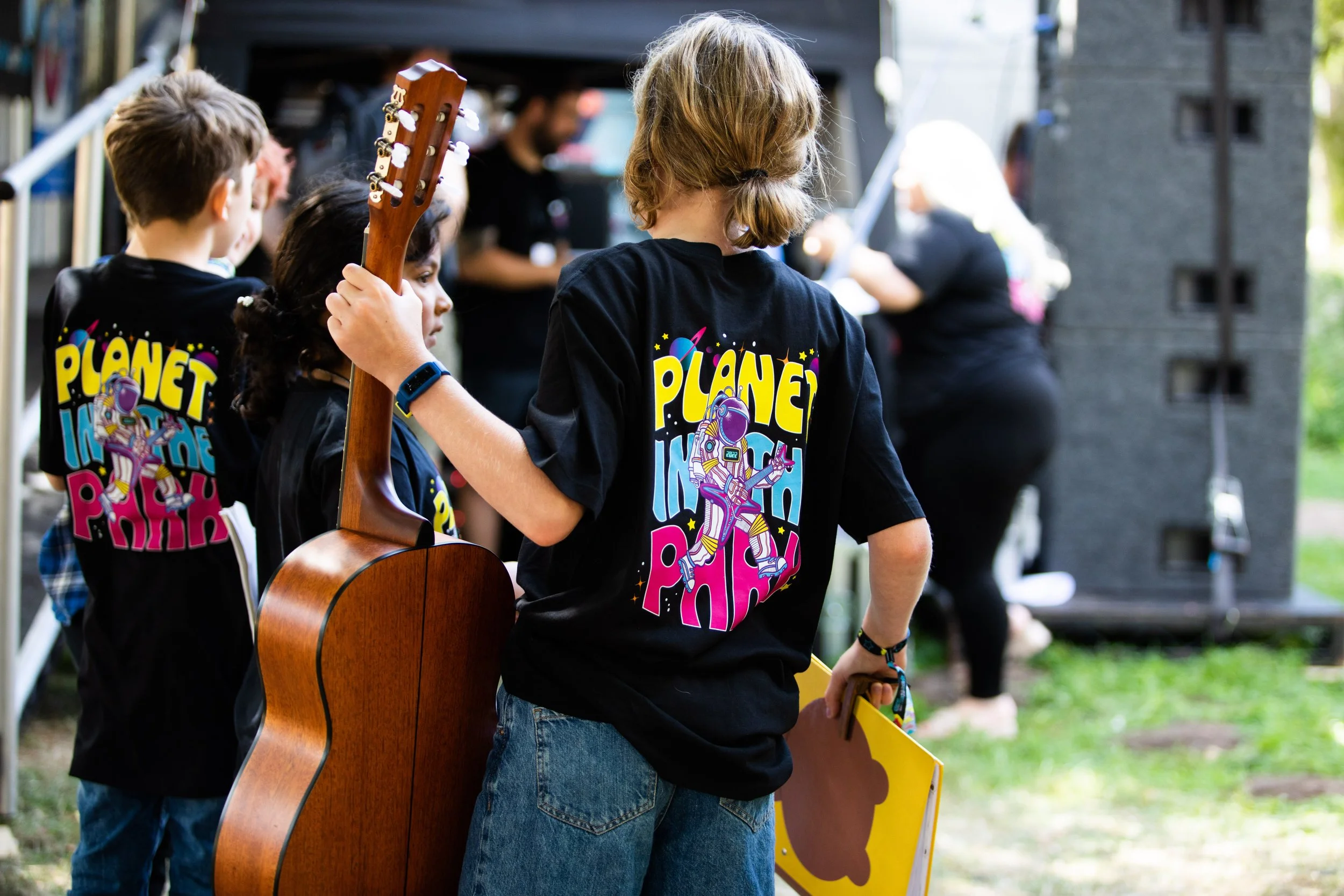 Children wearing matching and colorful 'Planet in the Pink' shirts, with one holding a guitar, standing outdoors near a stage with speakers, and adults standing in background.