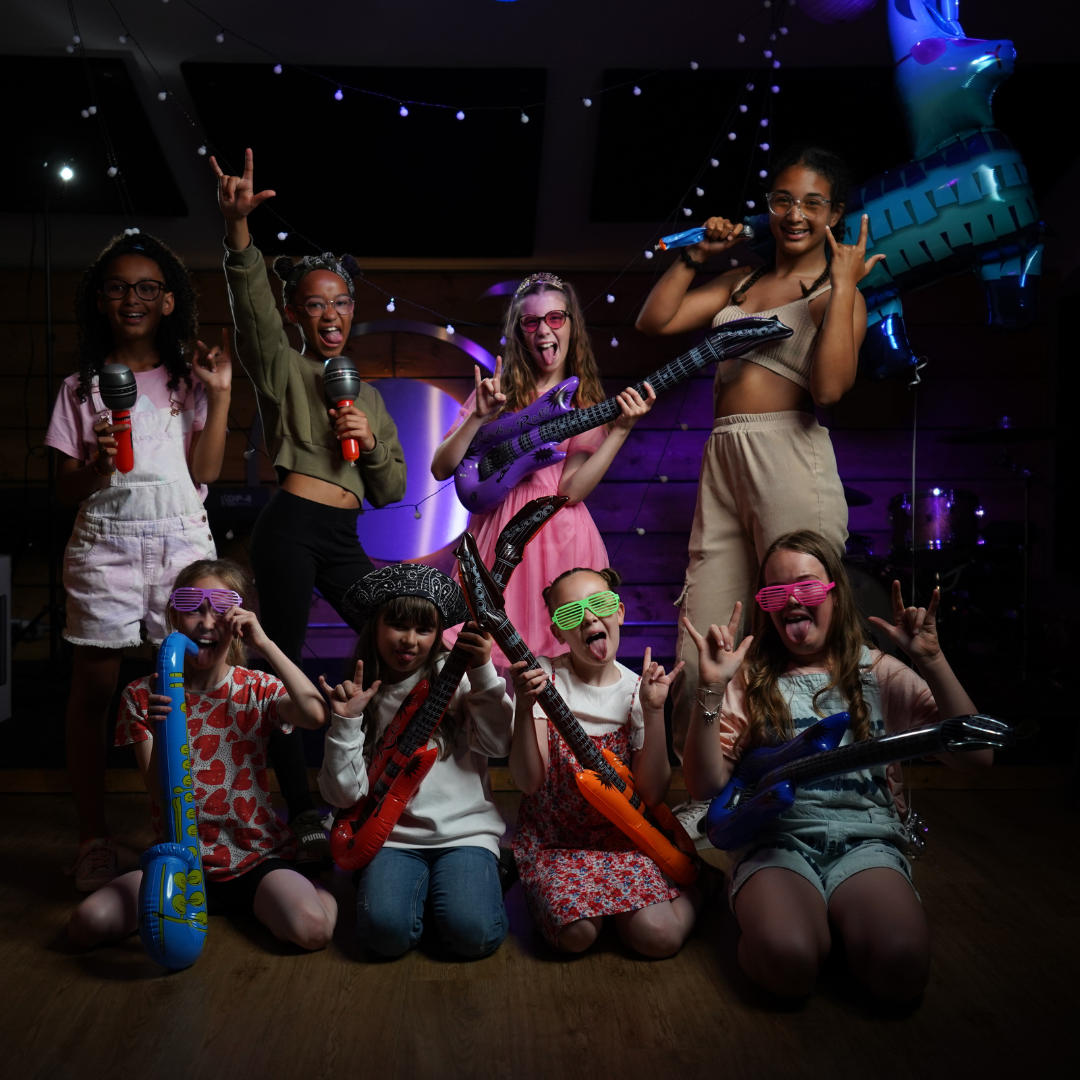 Children dressed in party attire at a birthday celebration, holding toy guitars and microphones, with colorful neon glasses, balloons, and festive decorations.