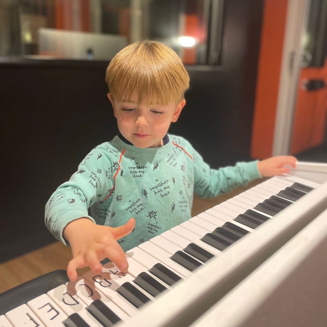 A young boy with red hair playing a white piano in a room with dark wall and window.