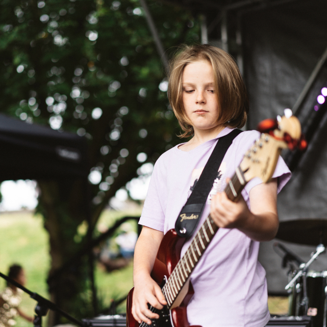 A young girl playing an electric guitar outdoors during daytime, standing in front of a blurred background of trees and people.