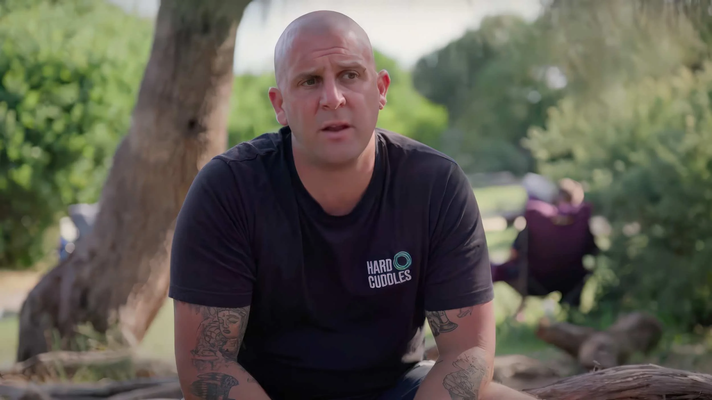 James Harding, wearing a black T-shirt with the words 'HARD CUDDLES' and a circular logo, sitting outdoors near a tree with a blurry background of people and greenery.