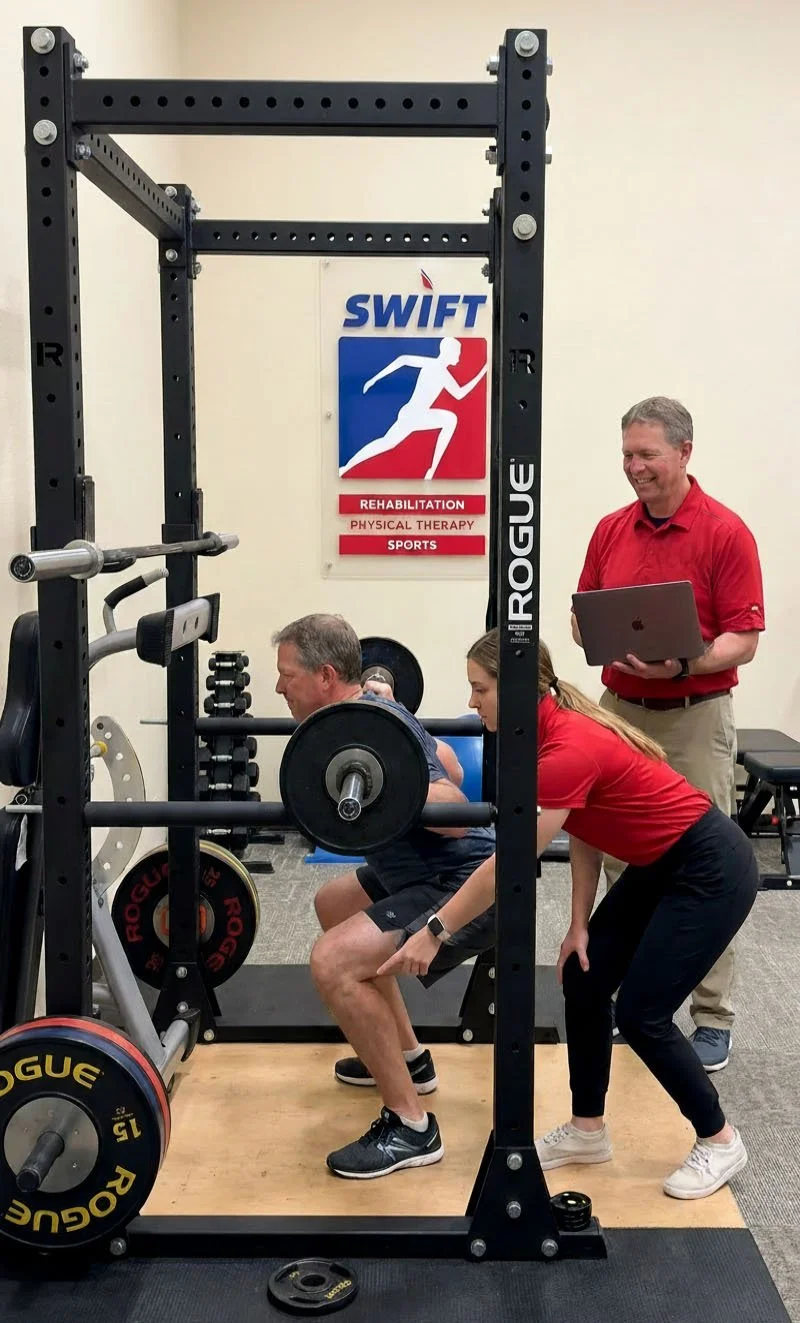 A man is performing a squat exercise with a barbell on his shoulders in a gym, supervised by a female trainer. A man in red shirt is standing nearby with a laptop, observing. There is a sign on the wall that reads 'SWIFT REHABILITATION PHYSICAL THERAPY SPORTS'.
