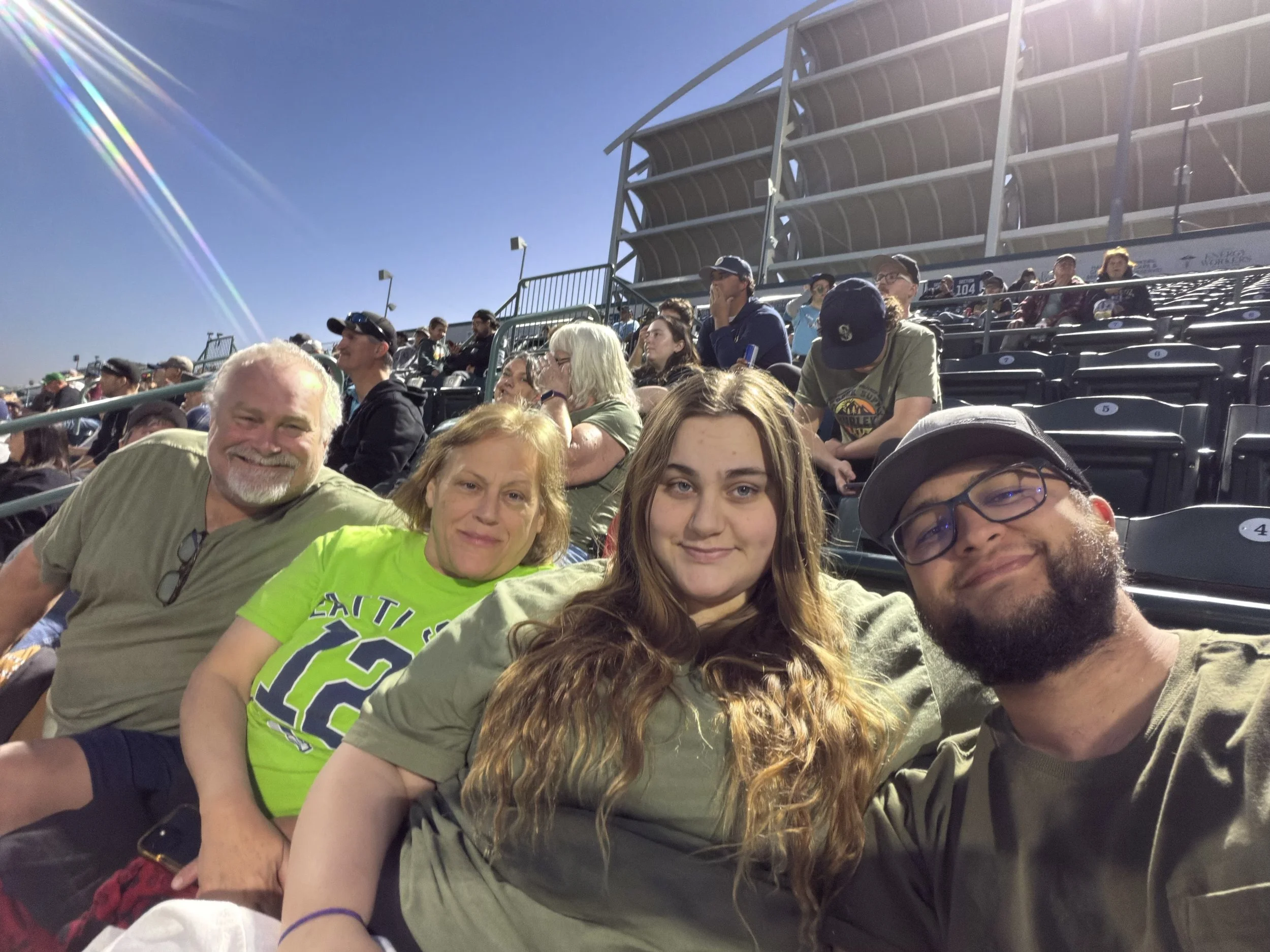 A group of people sitting in stadium seating on a sunny day, with a clear blue sky and bright sunlight causing lens flare.