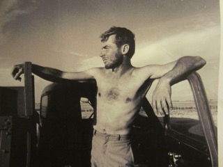 An old photograph of a young Richard, shirtless, leaning against the door of a pickup, gazing thoughtfully into the distance against an open sky.