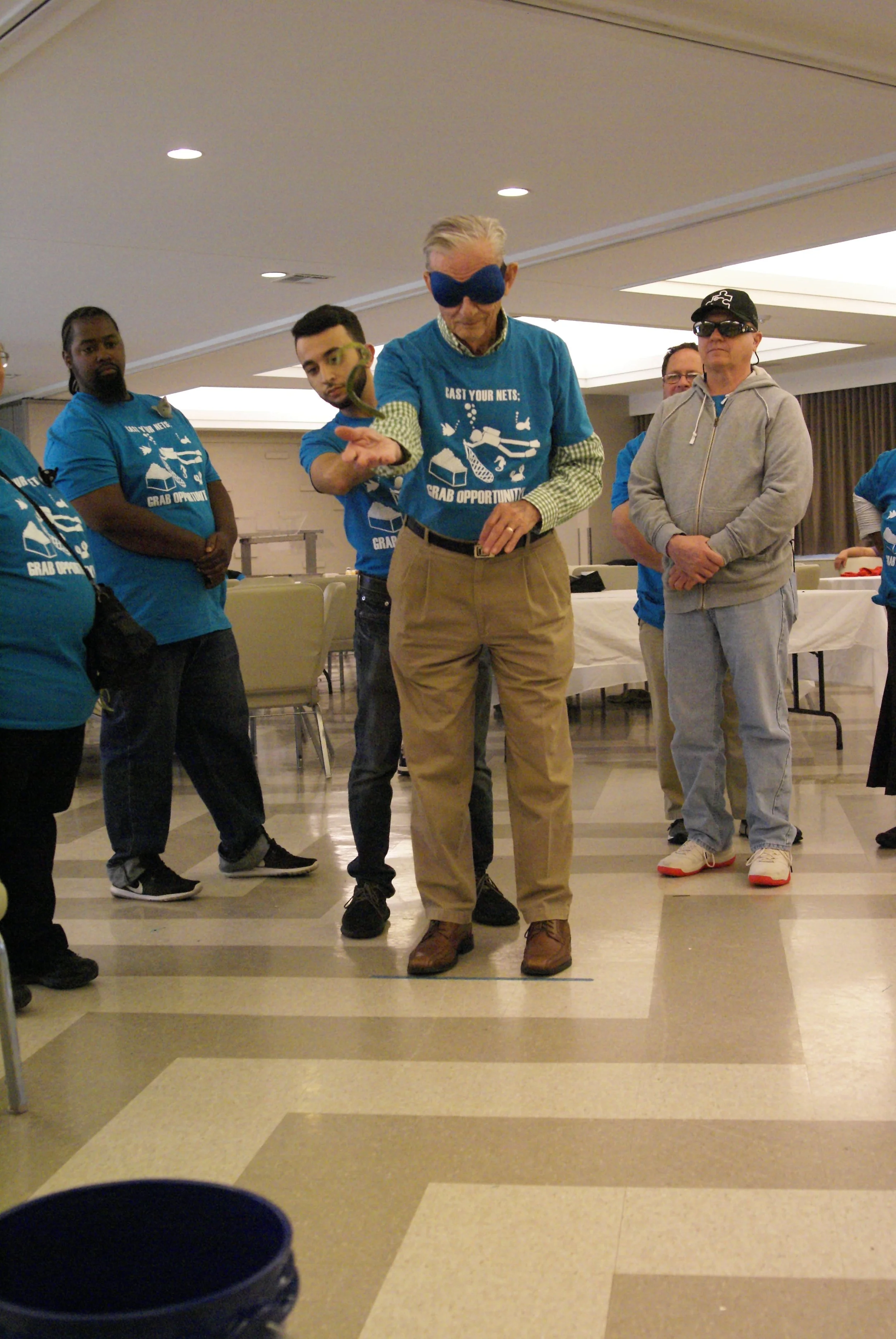  Picture Description: Deaf-Blind playing a toss game with the help of a SSP. The Deaf-Blind is wearing a blind fold. Two other SSPs standing watching the Deaf-Blind play the game  