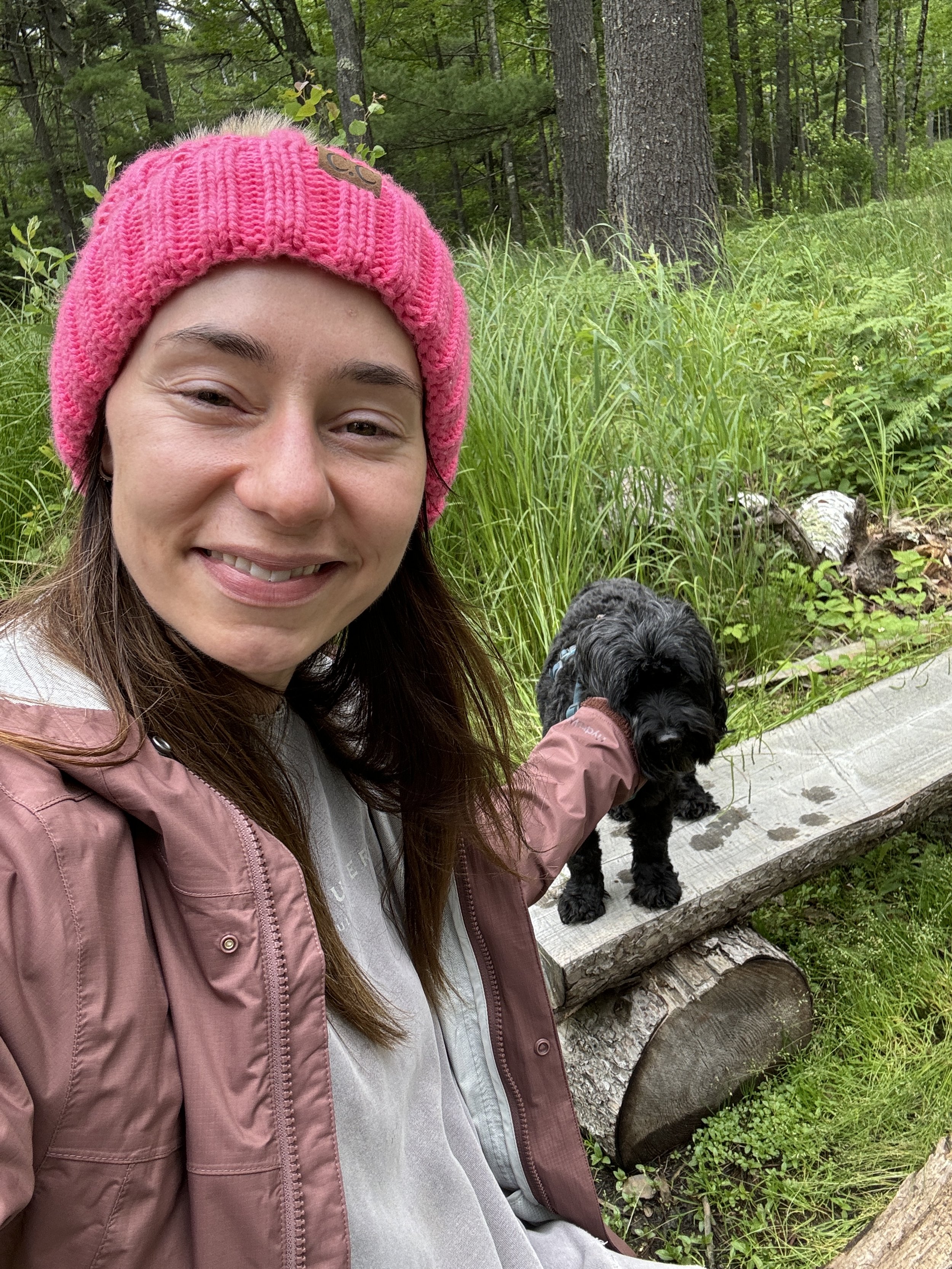 CrossFit Hopkins nutrition coach in a pink knit beanie and brown jacket smiling at the camera while holding a small black dog on a leash in a lush green forest.