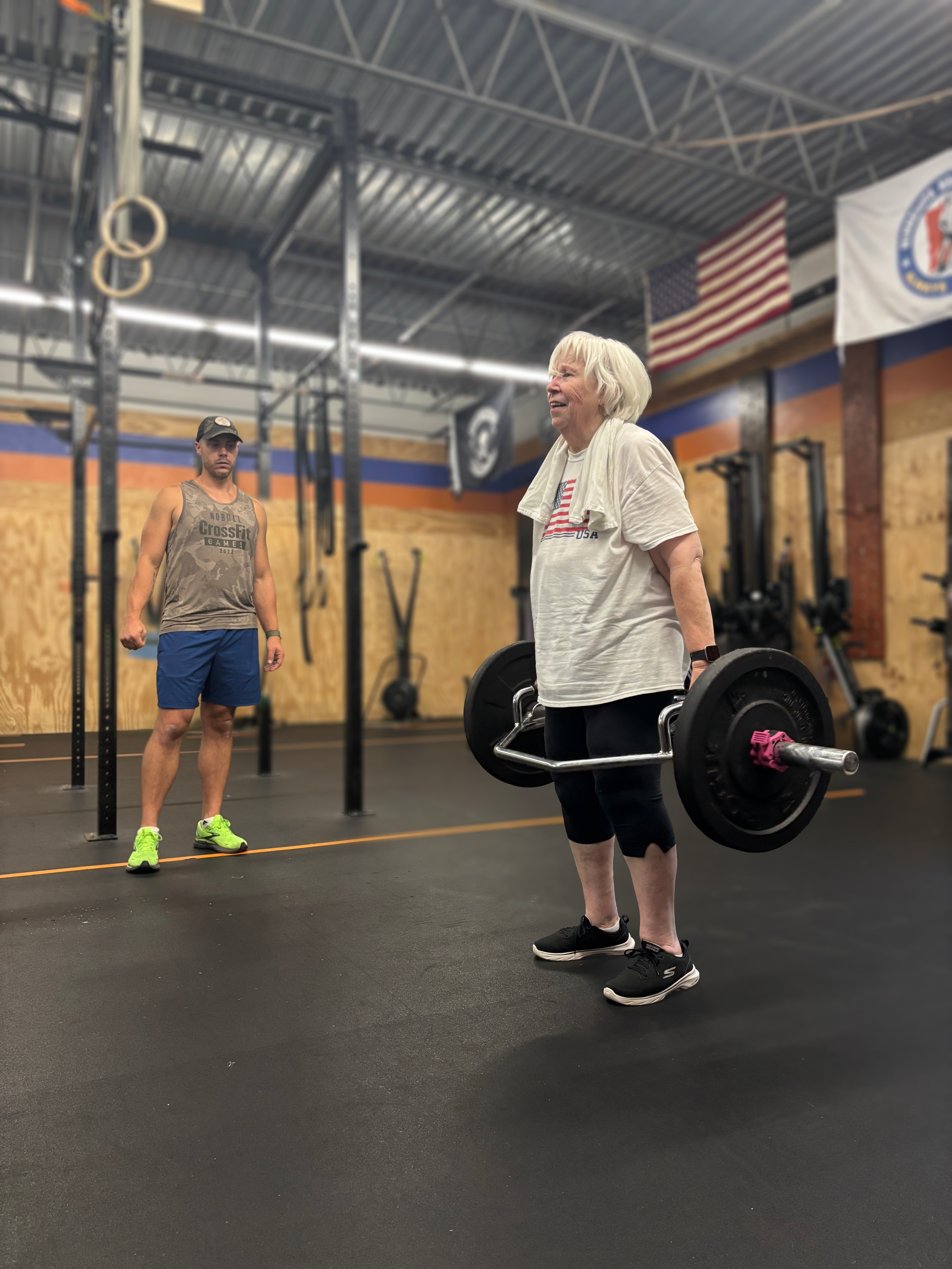 A senior woman in a white t-shirt lifting a barbell in a gym. A coach in a CrossFit tank top and shorts stands nearby, observing. The gym has a wooden wall, flags, and fitness equipment.