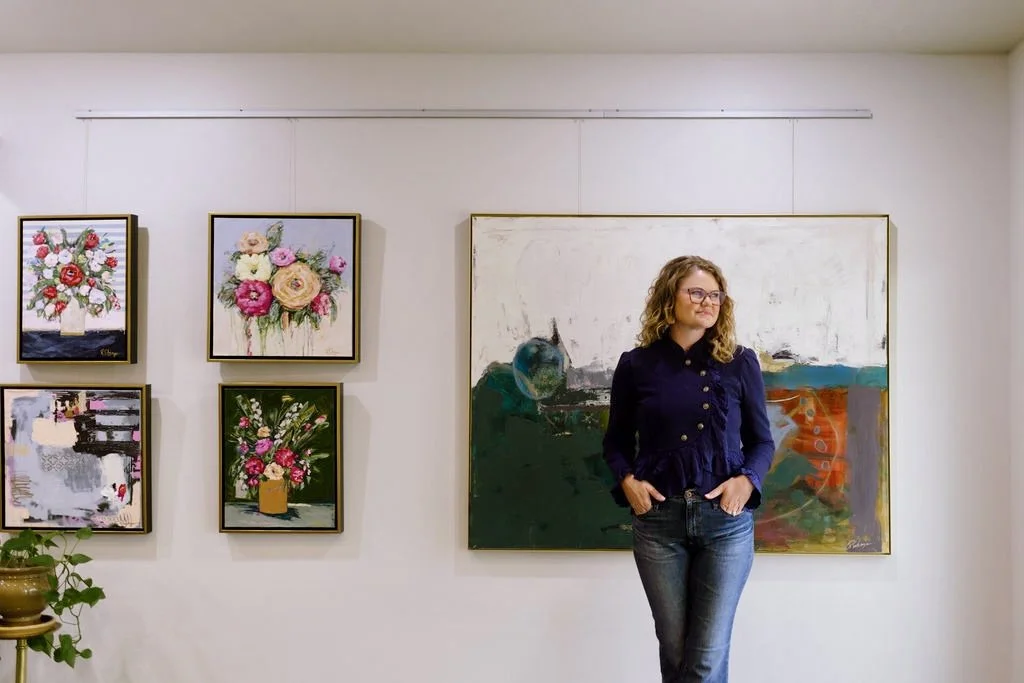 Artist posing in front of a mix of an large abstract art piece and medium size abstrats and florals in the new gallery space for Rhonda Schrage Art