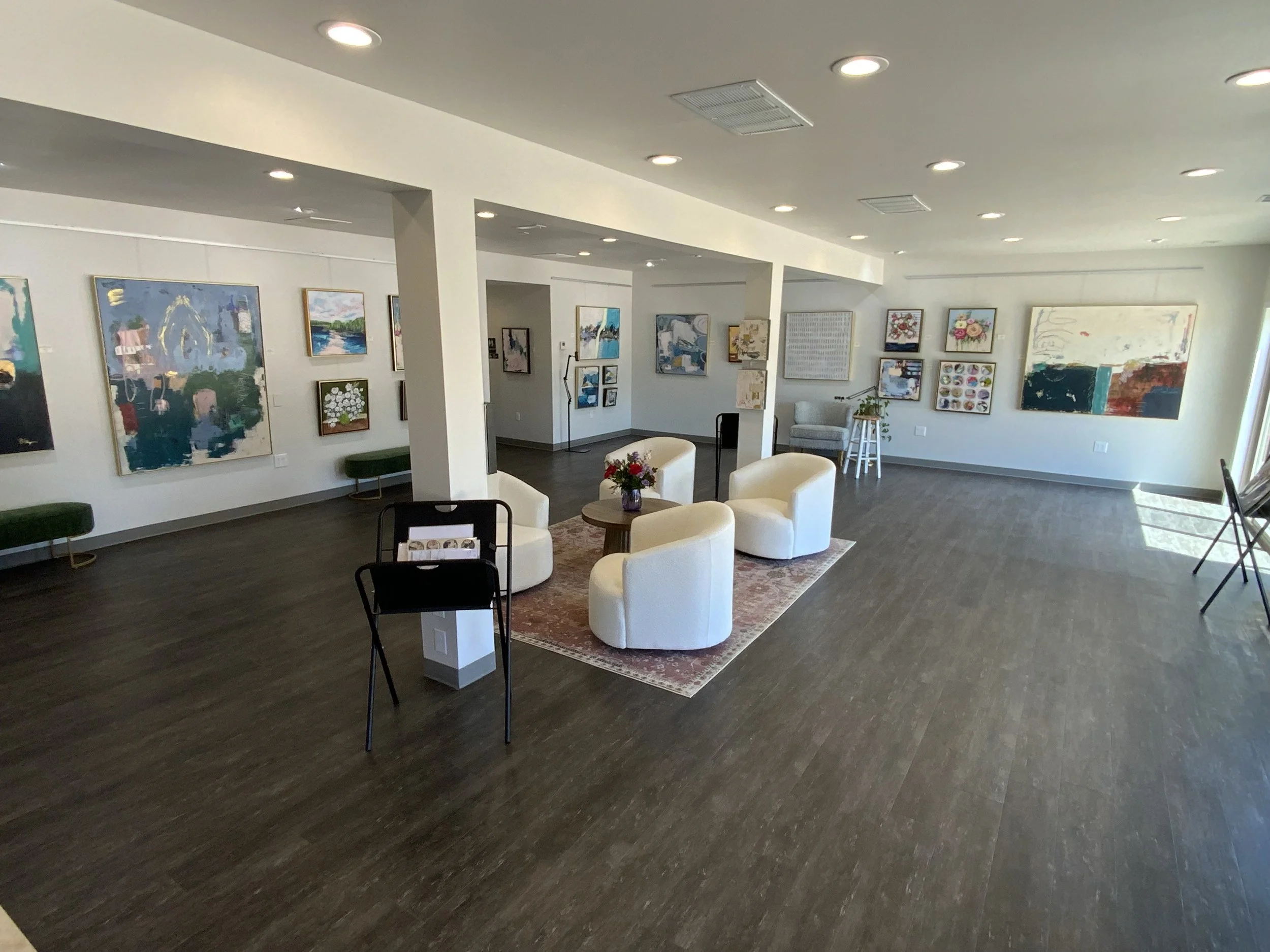 Image of the gallery space showing several pieces of abstract and floral art hung around the gallery with an inviting seating area in the center of the room. Rhonda Schrage Art gallery space.