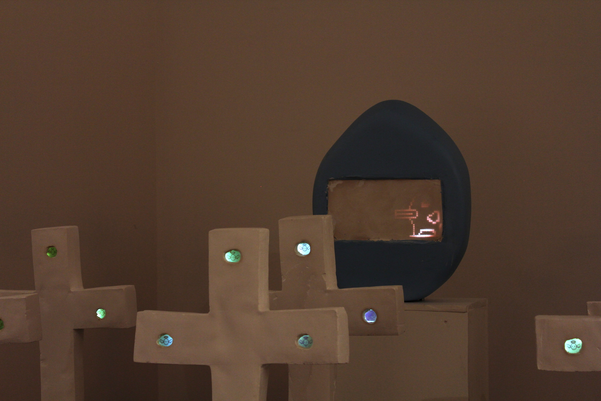 Four large plaster crosses glow with LCD screens with pixelated Tamagotchi-like angels. One large plaster egg features a duck-like Tamagotchi creater in a larger LED screen.