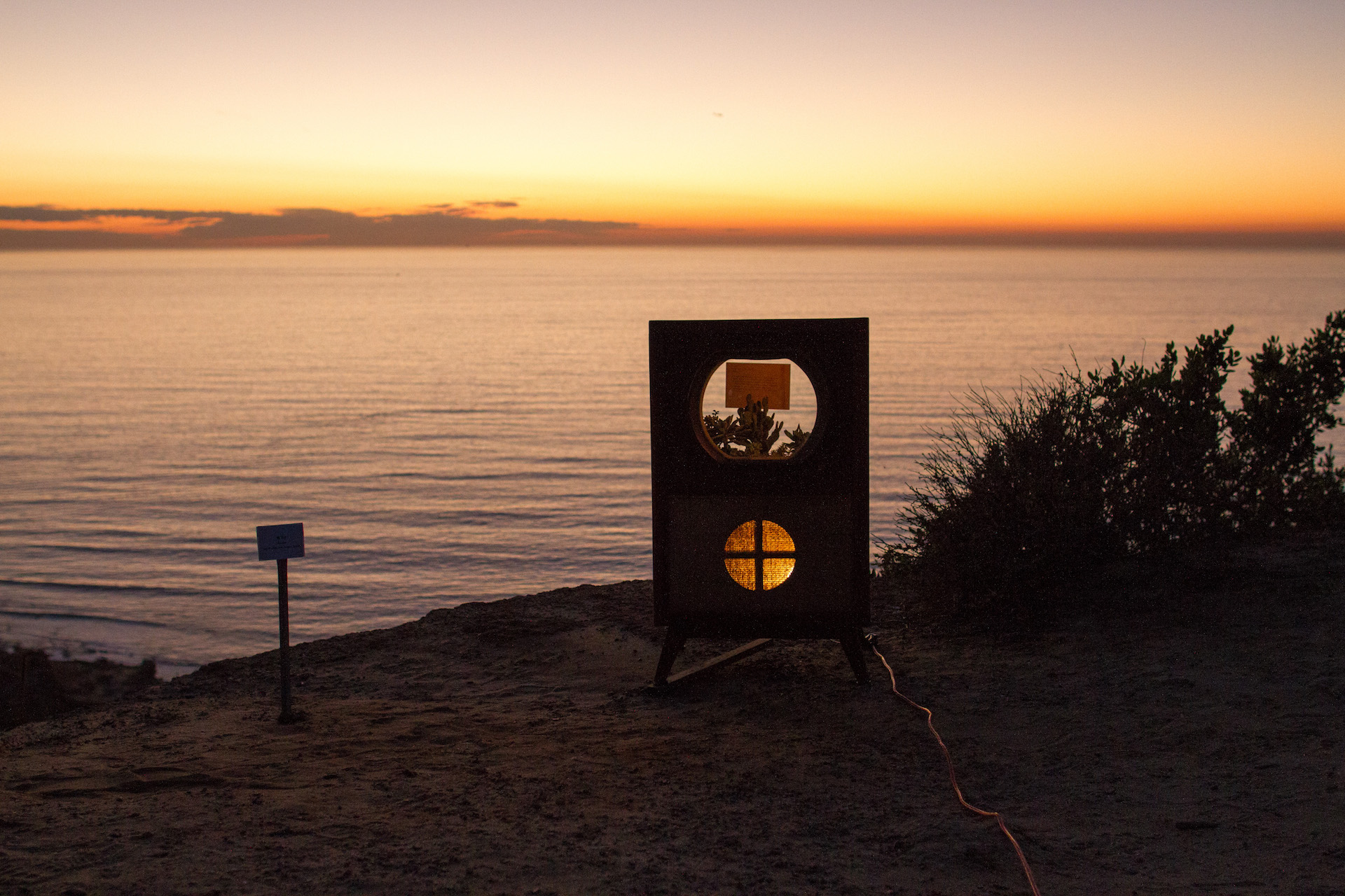 A silhouette of the tv set glows with an orange light at sunset. The same light reflects off of calm waves in the background.