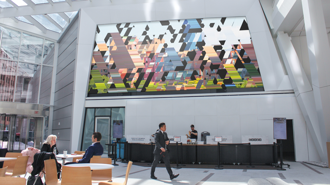 Business professionals hang out in an airy, sun-soaked lobby. A large LCD screen features a criss-crossing pattern of images and stretched pixels that looks like a deconstructed Boston neighborhood.