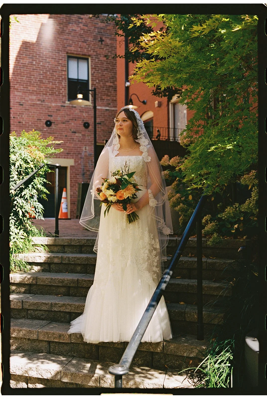 Bride in a white lace wedding gown and veil standing outdoors on stone steps, holding a bouquet of flowers, surrounded by greenery and brick buildings. Staunton Virginia Wedding Film Photographer.