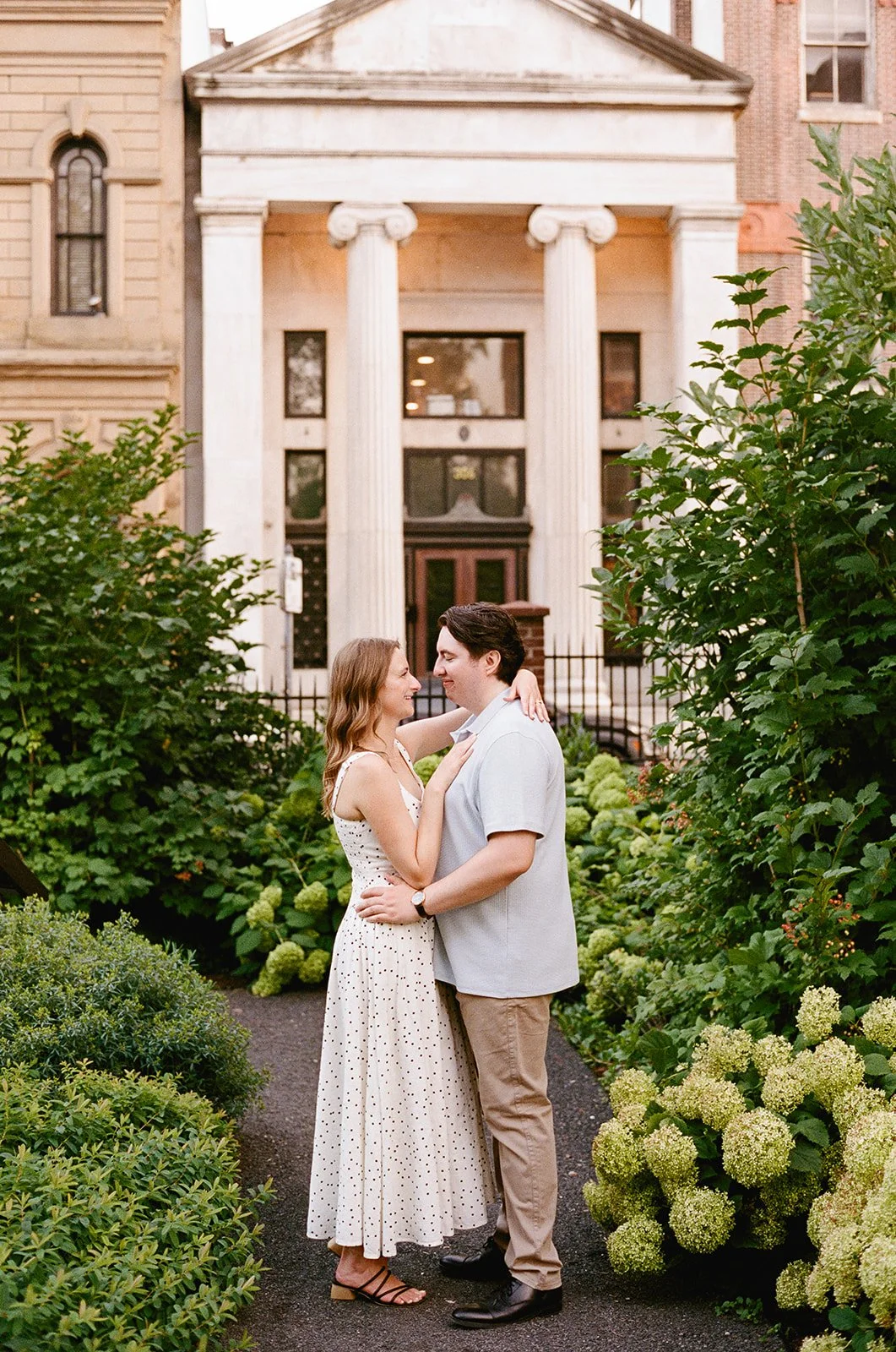 A couple standing close together in a garden in front of a historic building with white columns and a pediment, embracing and smiling. Old City Philadelphia engagement shoot