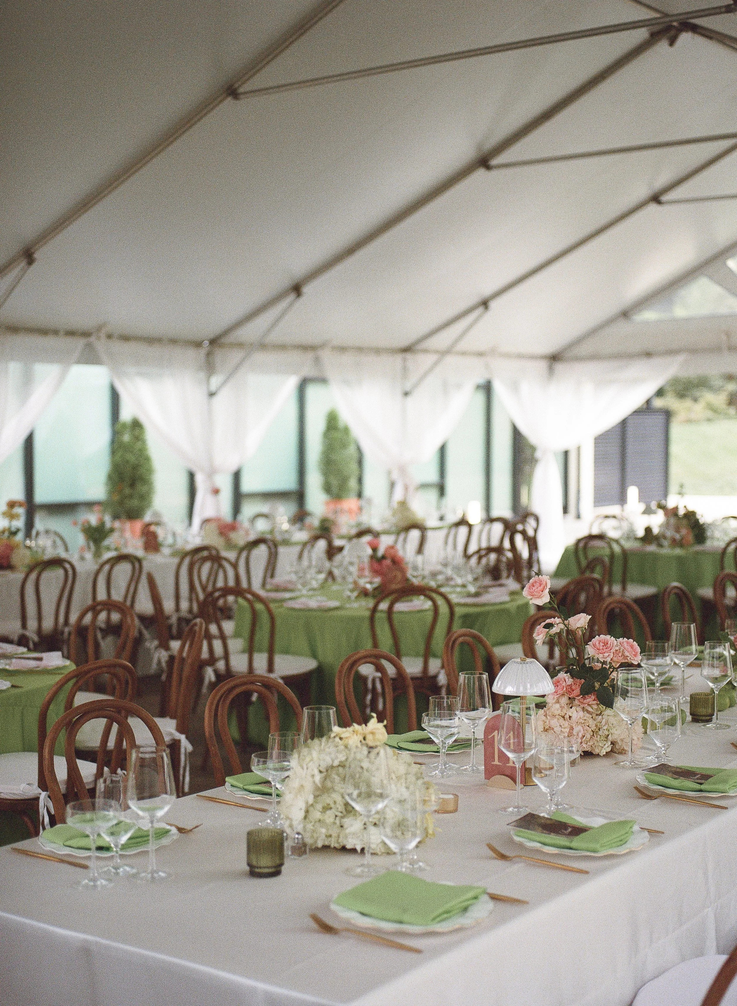 Indoor wedding reception setup with round tables covered with white tablecloths, decorated with pink and white floral centerpieces, green napkins, and glassware, under a tent with white curtains and a high ceiling.
