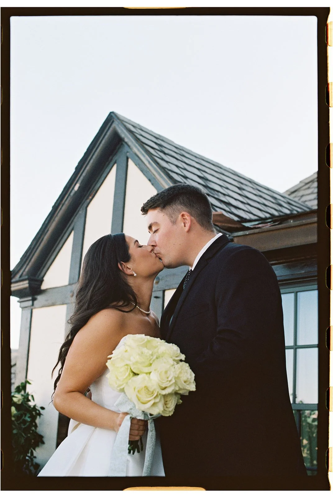 A newlywed couple sharing a wedding kiss outdoors, with the bride holding a bouquet of white roses, in front of a house with a steep roof and large windows. roanoke wedding photographer virginia