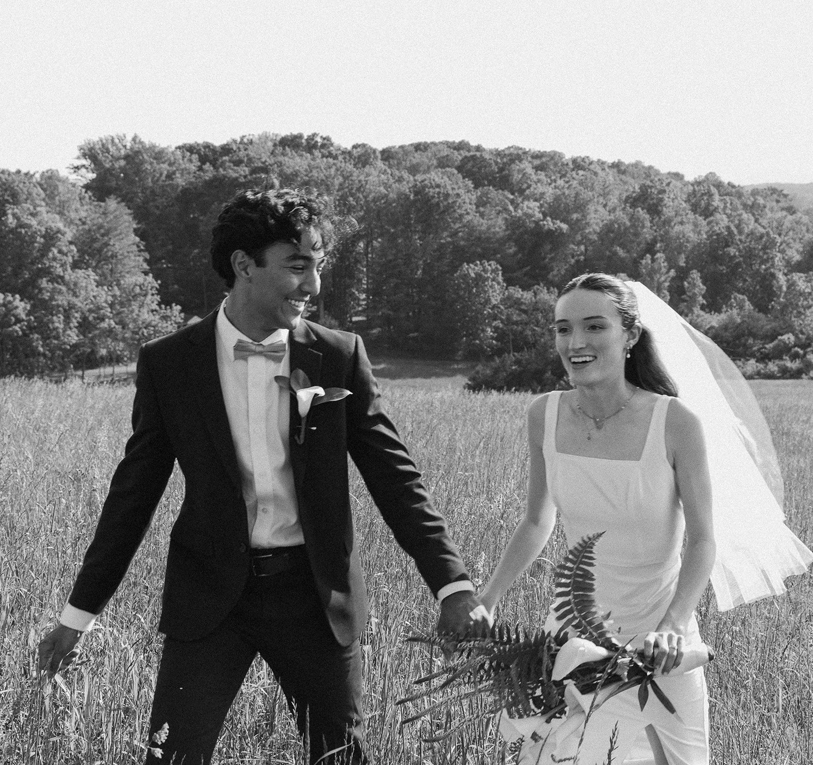 Black-and-white photo of a newlywed couple holding hands in a grassy field, smiling at each other, with trees and hills in the background. Lexington Virginia Wedding Film Photographer
