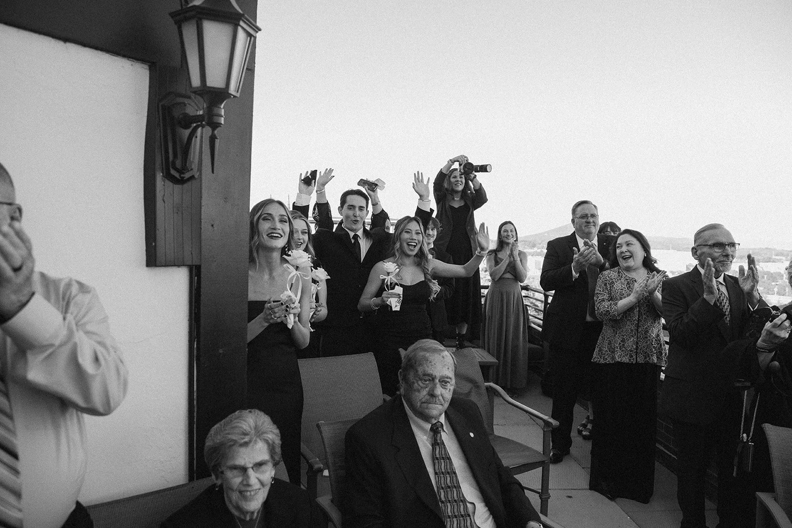 Black and white photo of a group of people celebrating and clapping on a balcony or terrace. Some are standing, some are seated, with a mix of formal and semi-formal attire. Several people are smiling and cheerful, and a woman is filming with a camera. A man in the foreground appears serious, while others look happy and excited.