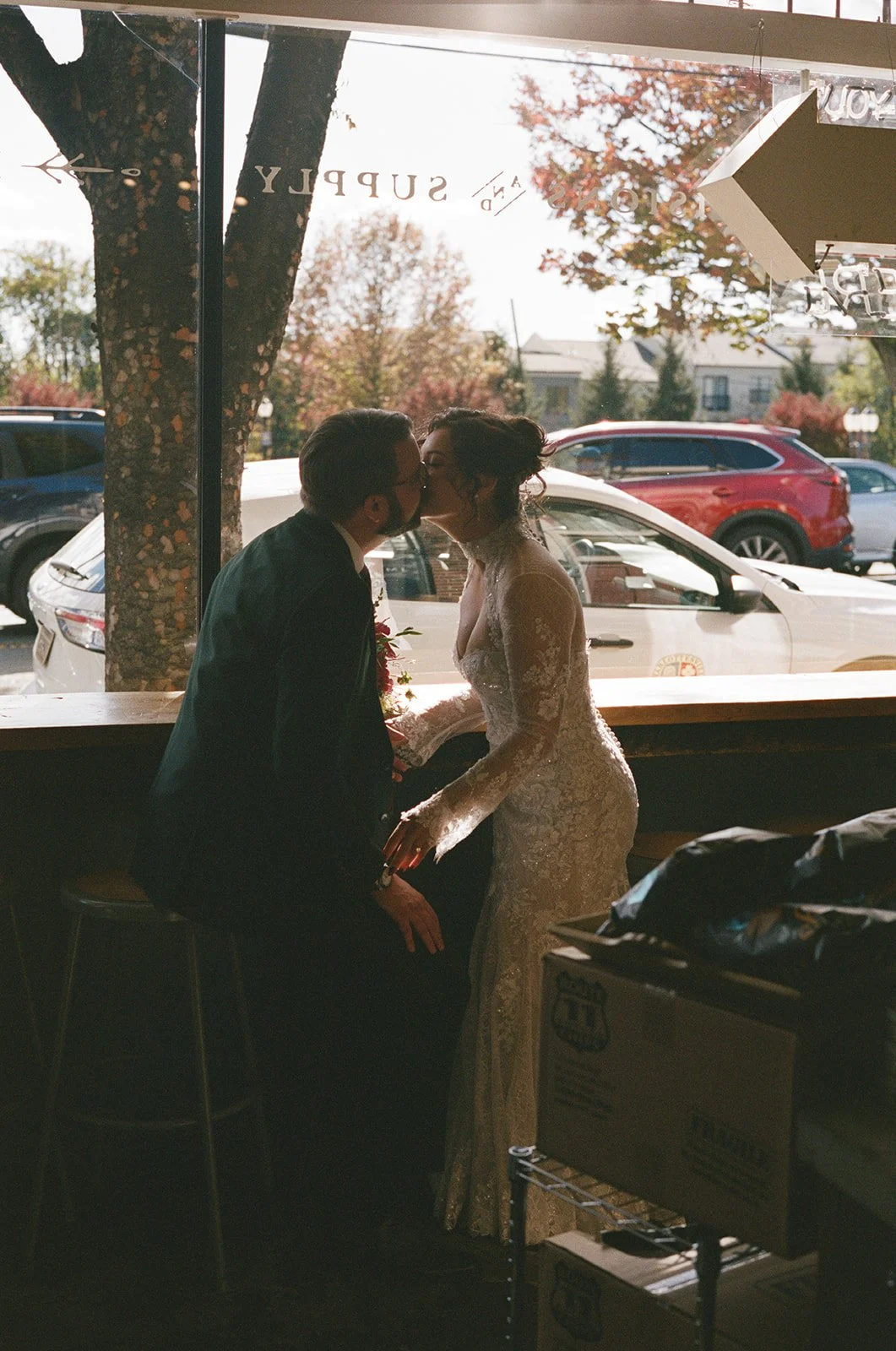 A couple dressed in wedding attire kissing inside a restaurant with a window showing parked cars and trees outside. downtown charlottesville va