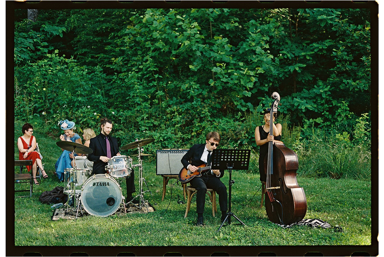 A group of musicians performing outdoors on a grassy area with lush green trees in the background. The band includes a drummer, a guitarist, a bassist, and a keyboardist, with two women sitting in the background observing the performance