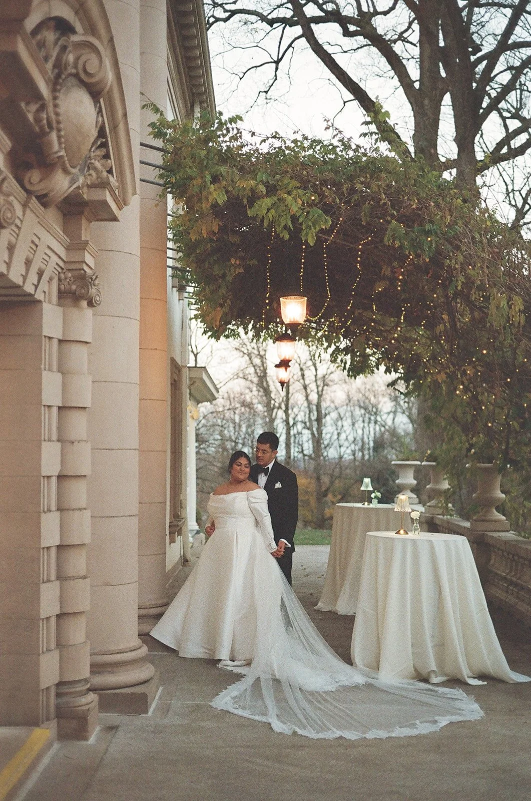 A bride in a white wedding gown and a groom in a black tuxedo holding hands, standing outside under hanging lights and string lights, with tables covered in white cloth and small lamps, trees, and cloudy sky in the background. Liriodendron Mansion
