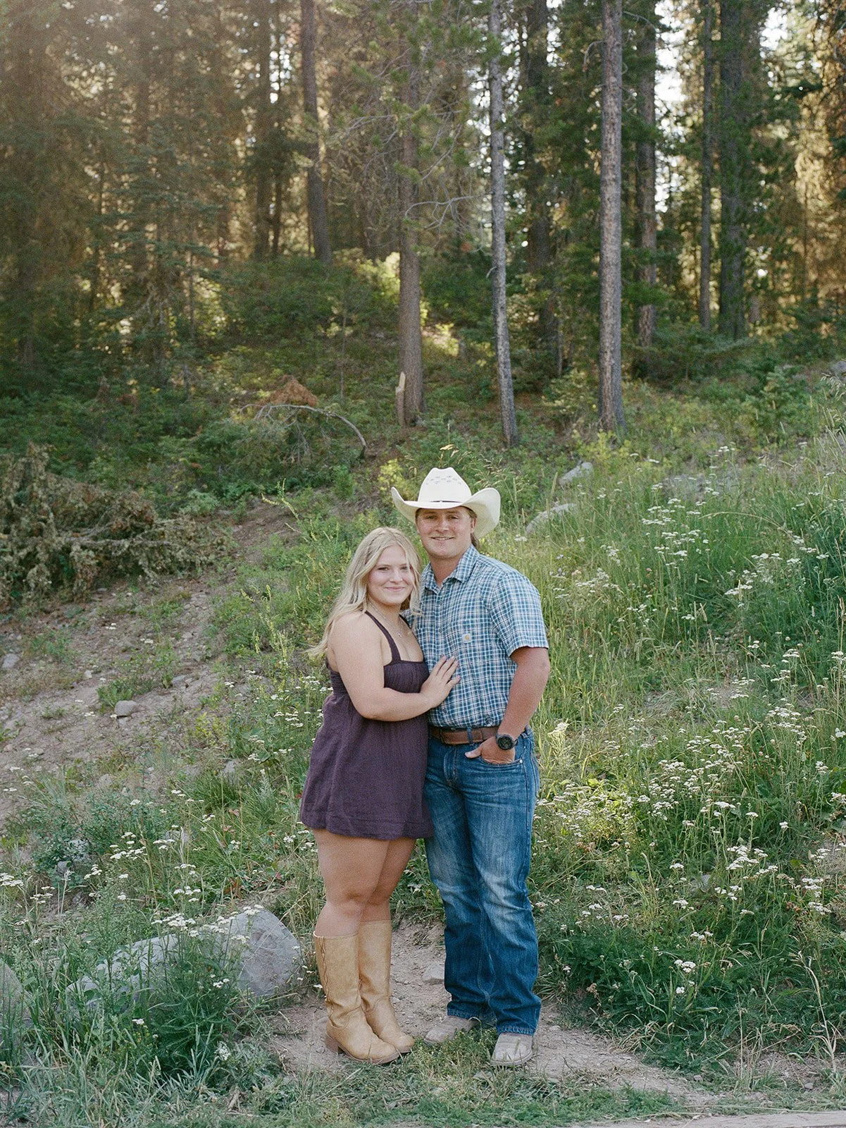A couple standing outdoors in nature with tall grass and wildflowers, surrounded by trees and a wooded forest background. The woman is wearing a dark purple dress and tan cowboy boots, and the man is dressed in a plaid shirt, jeans, and a white cowbo