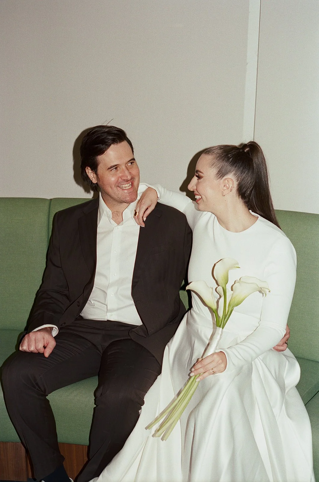 A smiling couple sitting on a green couch, the woman holding a bouquet of white calla lilies, the man in a black suit and white shirt, both laughing and looking at each other. nyc wedding film photographer