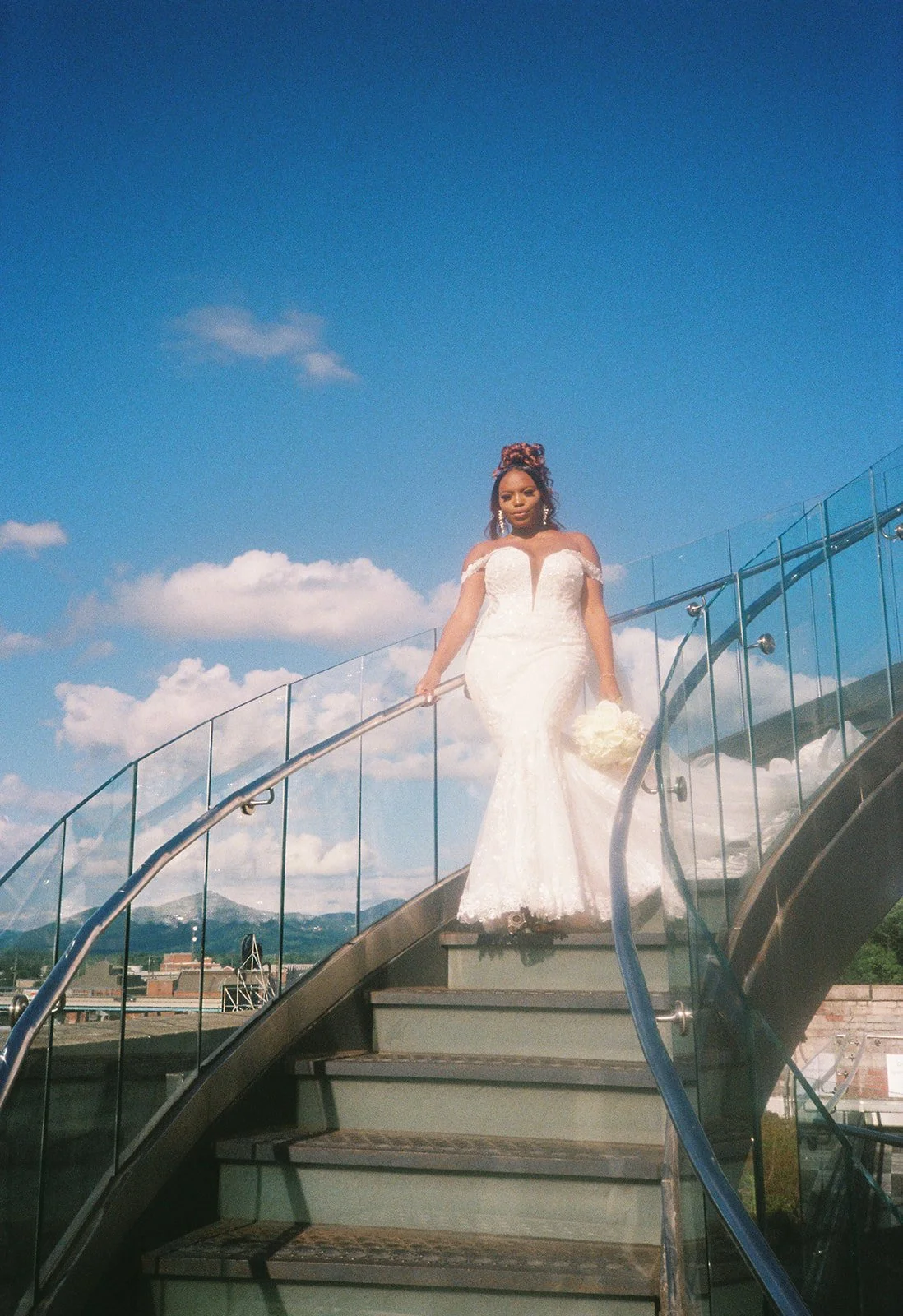 Bride in a white wedding dress ascending an outdoor glass staircase against a blue sky with clouds, holding a bouquet of white flowers in Roanoke Virginia.