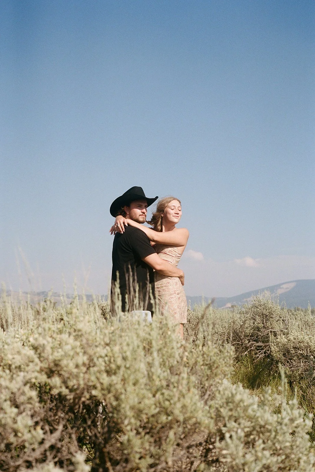 A man wearing a cowboy hat and a woman in a strapless dress embrace in a field of dry plants with mountains in the background on a clear, sunny day. Mormon Row Jackson Hole Wyoming engagement photos