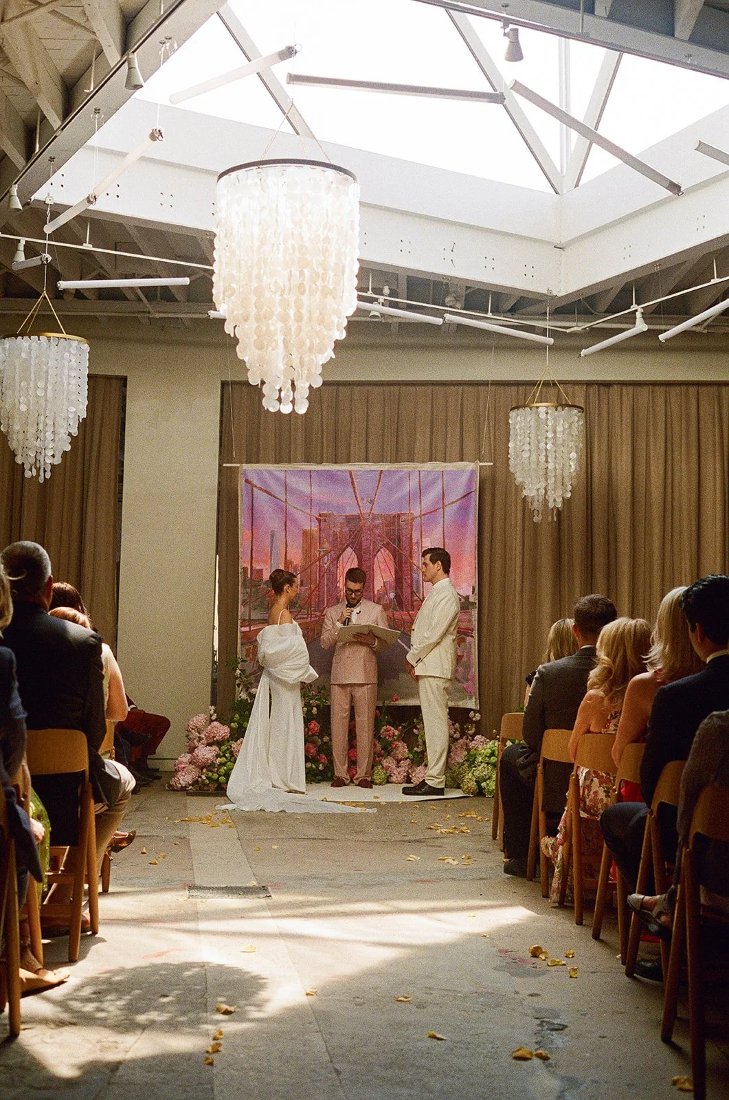 A wedding ceremony taking place indoors under a large skylight, with guests seated on either side. The couple stands at the altar with a officiant, and a backdrop of the Brooklyn Bridge is behind them. Floral arrangements and chandeliers decorate the room.