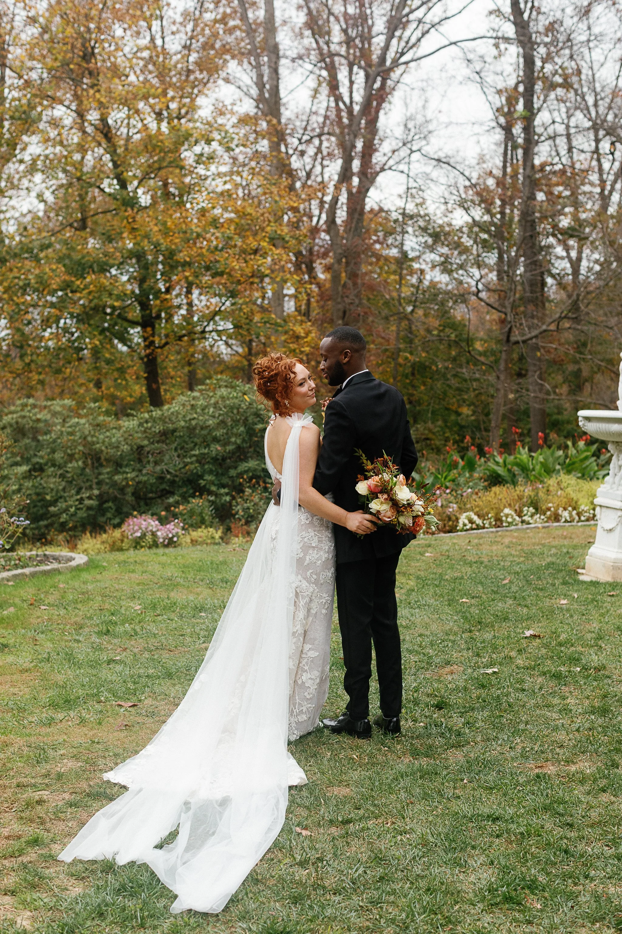 Bride and groom outside in a garden, embracing romantically. Bride in a white gown with a long veil, groom in a black suit holding a bouquet. gramercy mansion maryland wedding