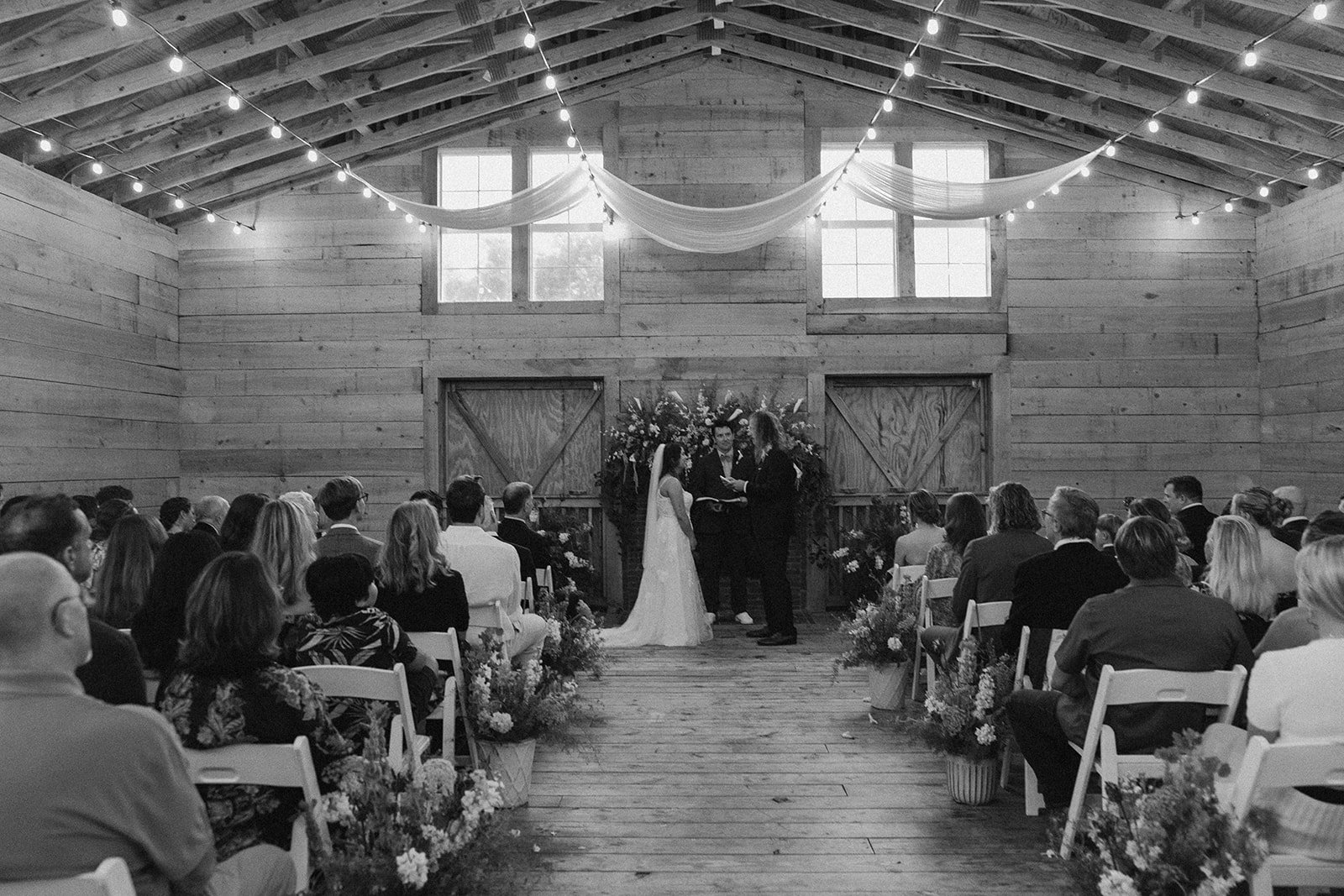 A wedding ceremony taking place inside a rustic wooden barn with guests seated on either side, a bride and groom standing before a minister, decorated with flowers and string lights. ocracoke outerbanks wedding film photographer