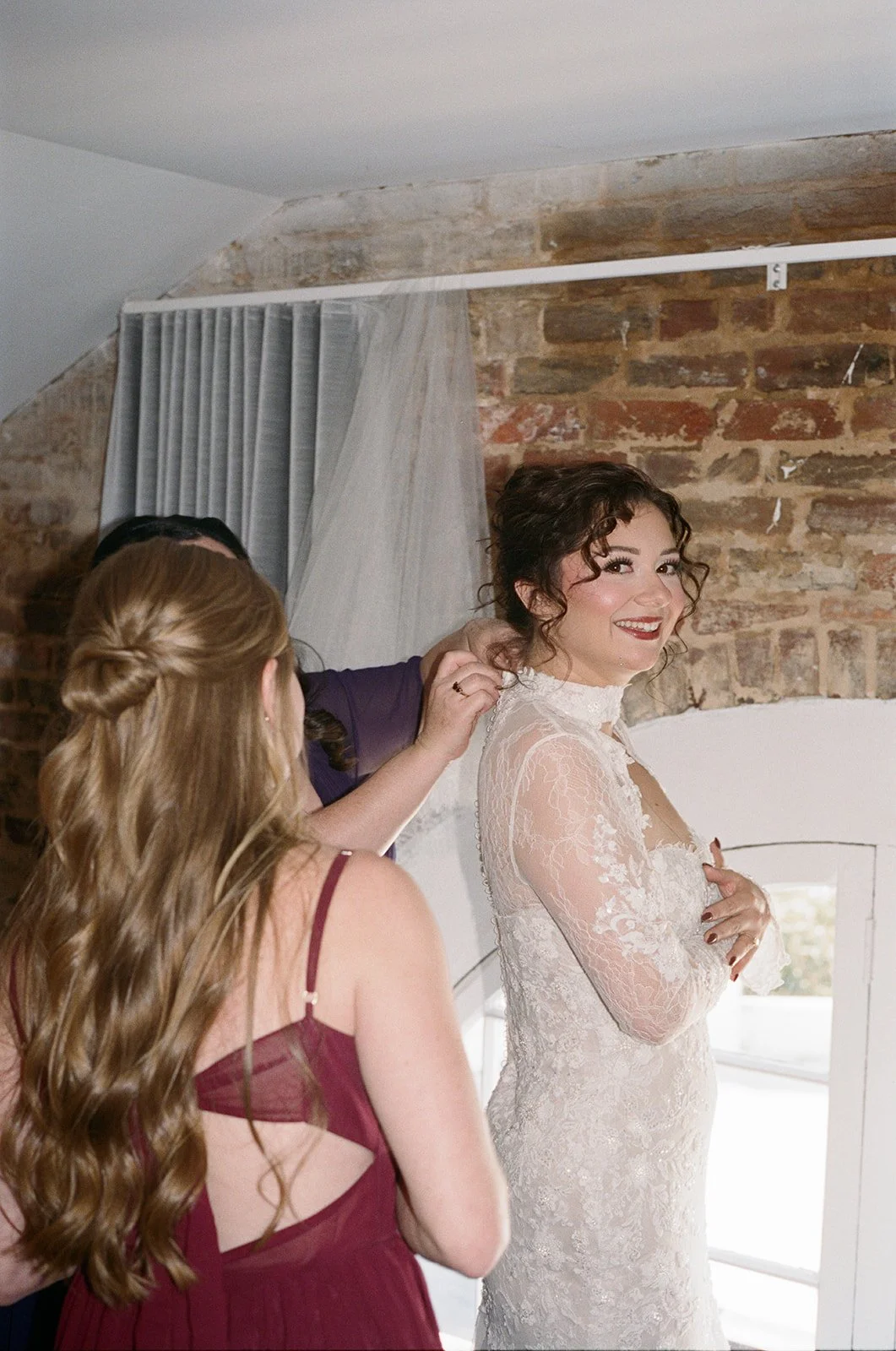 A bride in a lace wedding dress smiling as someone helps her with her hair, with another woman in a burgundy dress in the foreground, in a rustic room with brick walls and a window. doyle hotel charlottesville va