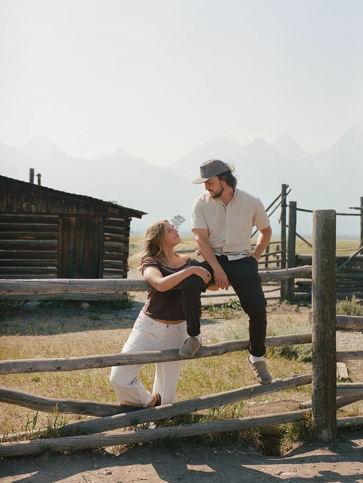 A man and woman sitting and leaning on a wooden fence outdoors, with a rustic cabin and mountain landscape in the background. mormon row jackson wyoming engagement shot