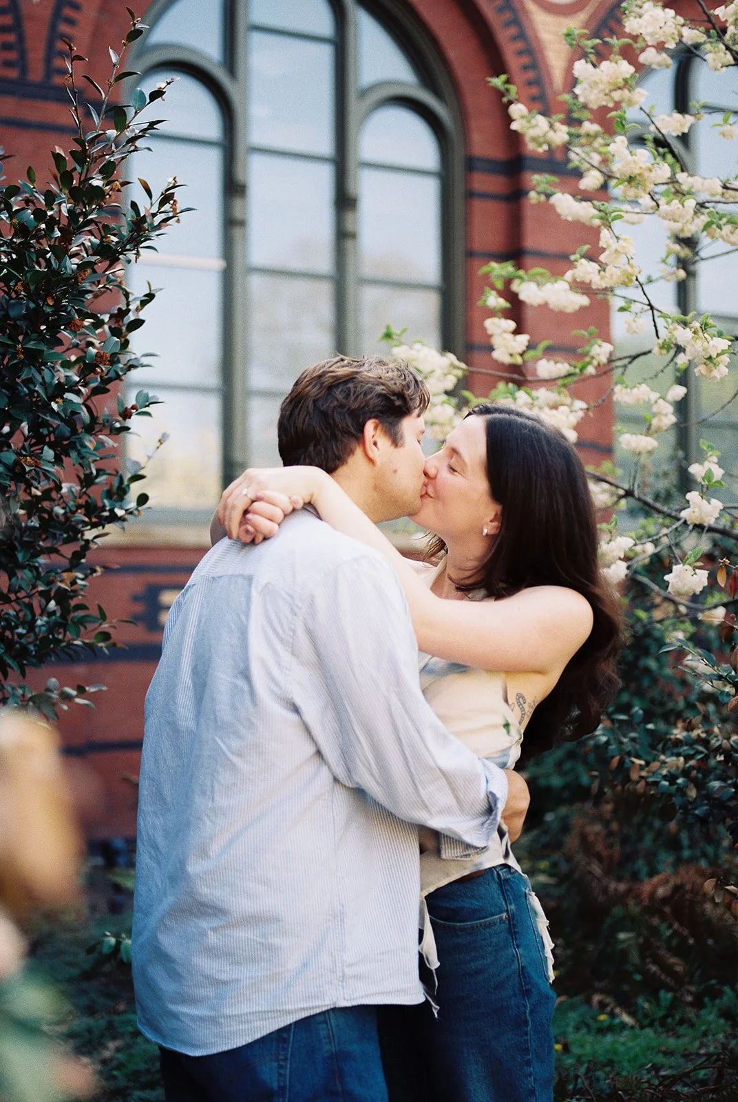 A man and woman sharing a kiss outdoors near blooming white flowers and a brick building with large arched windows.