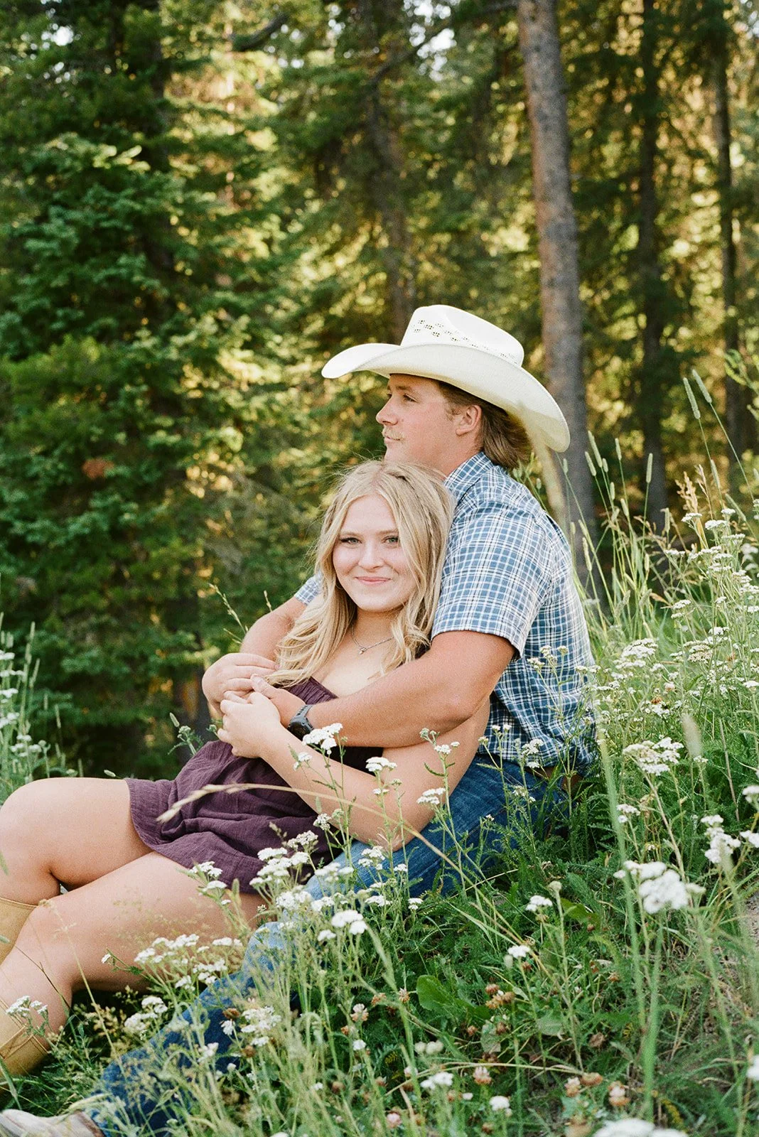 A young man wearing a cowboy hat and plaid shirt sitting in a field of white wildflowers, holding a young woman with long blonde hair. Hyalite Reservoir Bozeman Montana