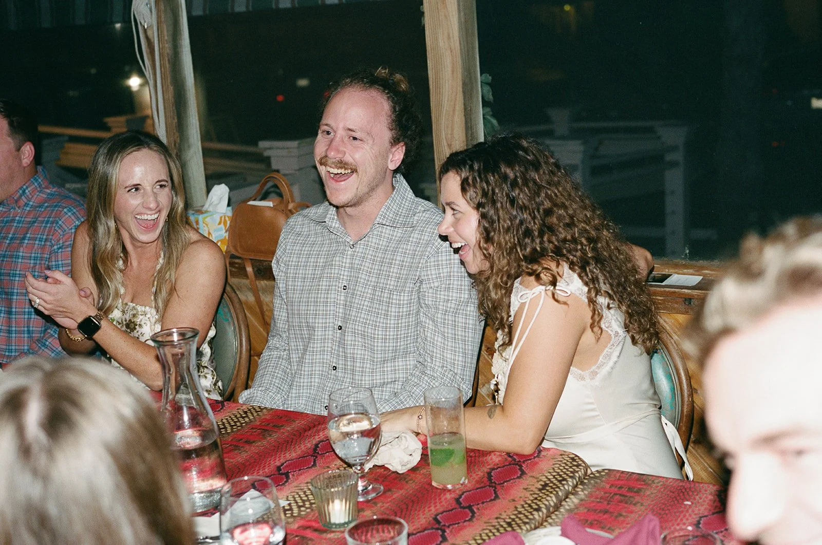 People laughing and enjoying themselves at a dinner party, sitting around a table with drinks, in a cozy indoor setting. ocracoke outerbanks wedding film photographer