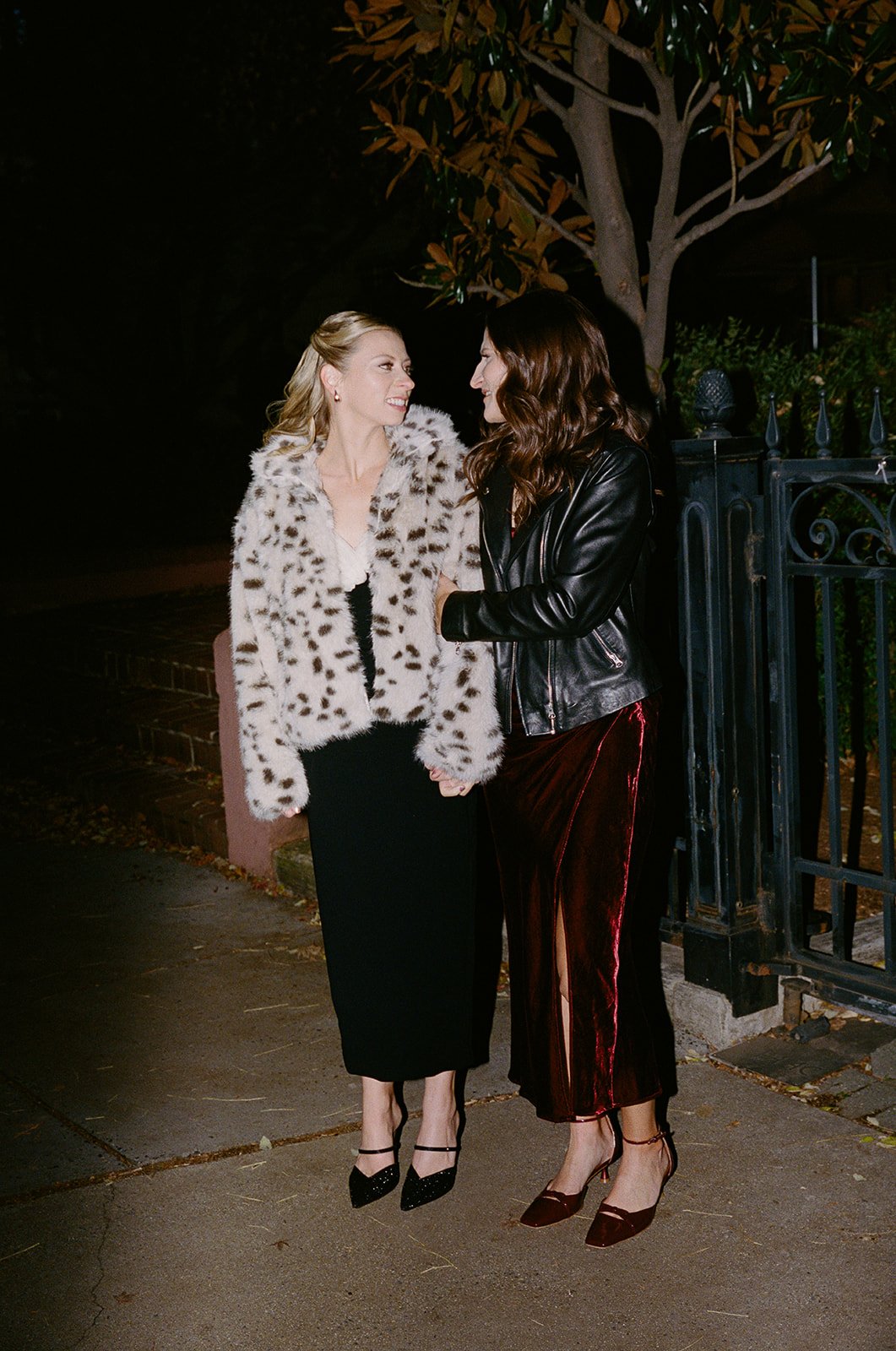 Two women standing outdoors at night, smiling and holding hands. One is wearing a leopard-print fur coat and black skirt; the other wears a black leather jacket and deep red velvet skirt. Washington DC wedding 35mm film