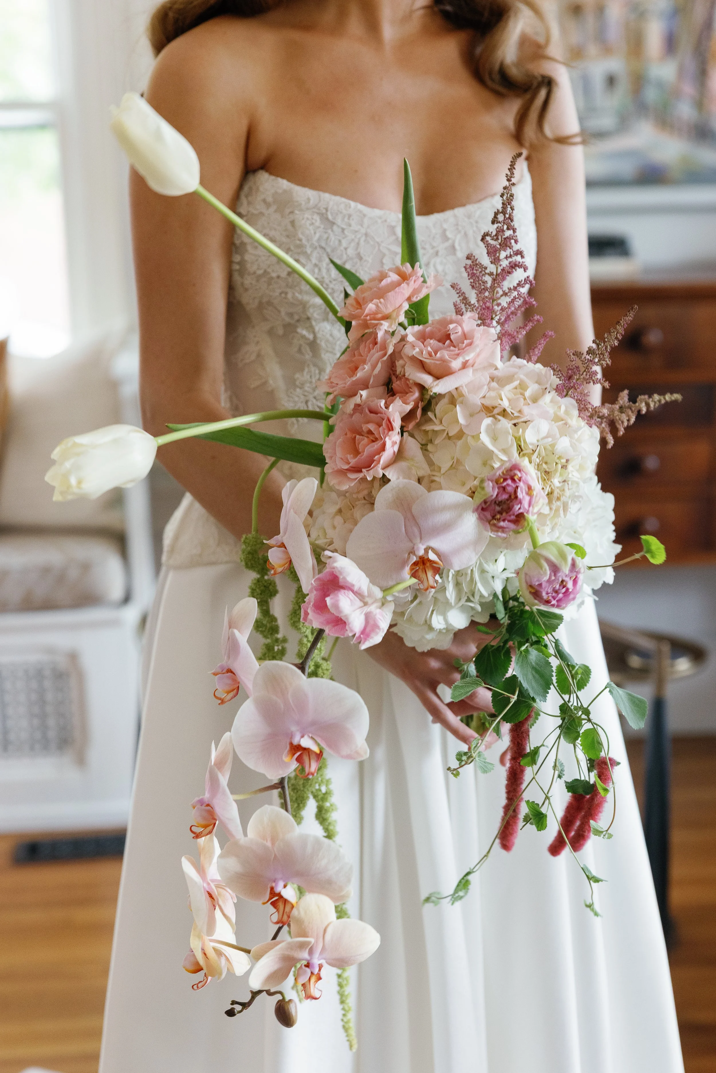 A bride in a strapless lace dress holding a cascading bouquet of white and pink flowers, including orchids, roses, and greenery, in a sunlit room. richmond virginia wedding photographer