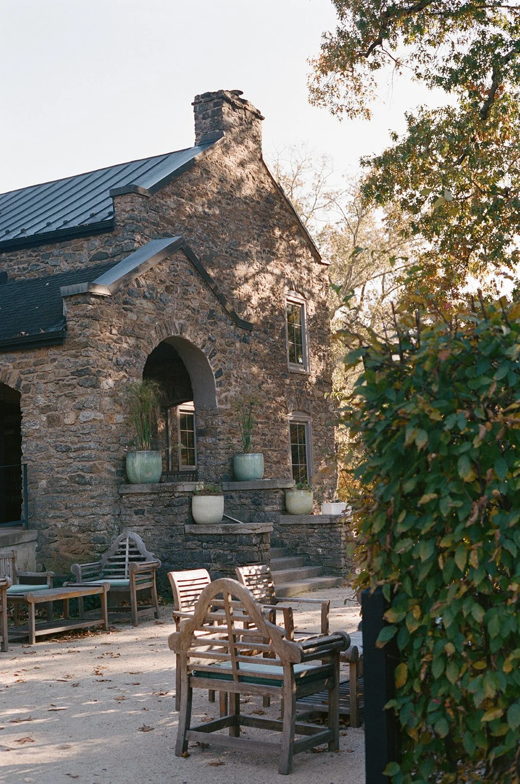 A stone house with a metal roof, arched window, and potted plants on the porch, surrounded by outdoor seating and greenery, in warm sunlight. potters craft cider charlottesville va
