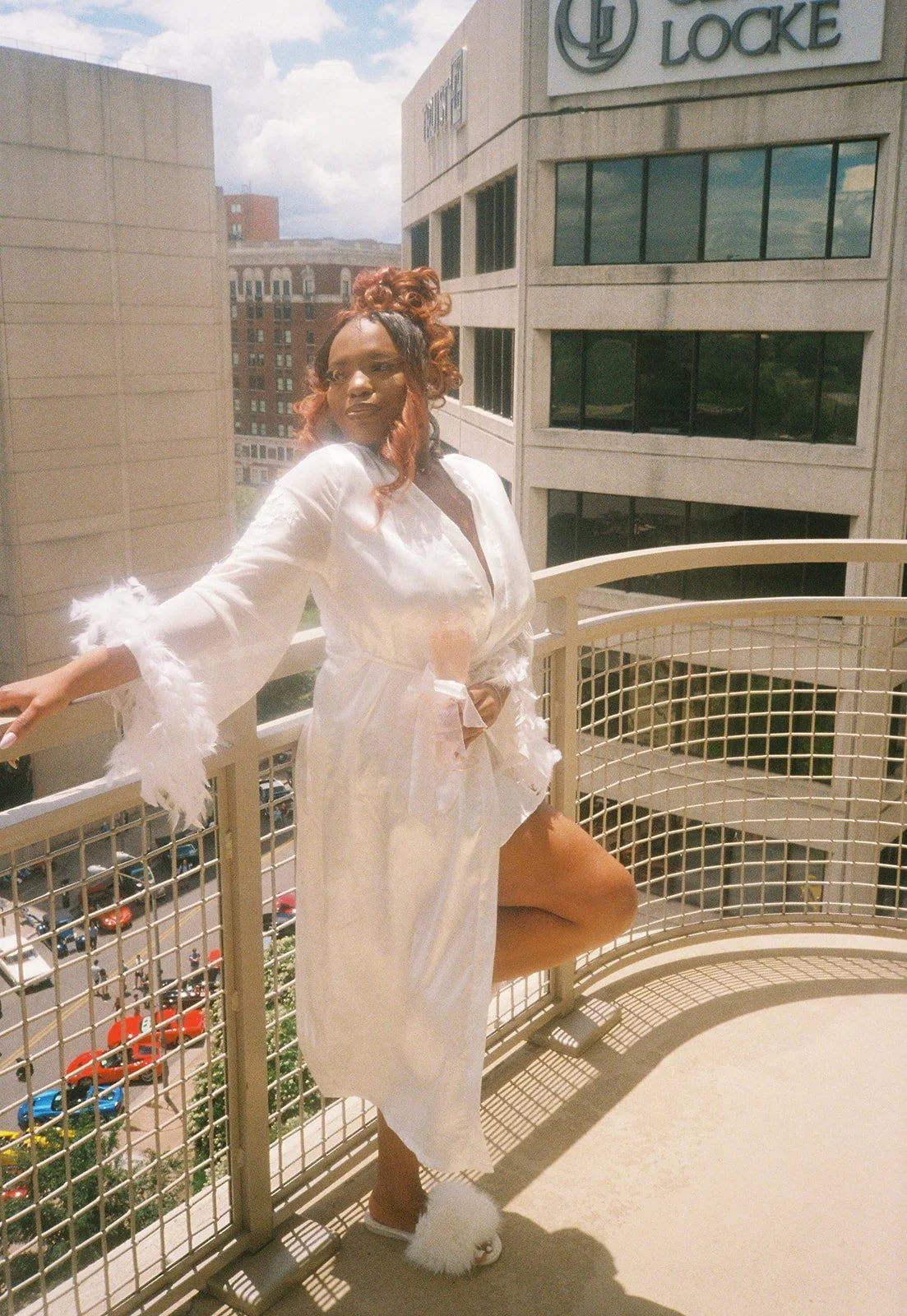 A woman in a white robe with feathered cuffs and slippers standing on a balcony with city buildings in the background. roanoke wedding film photographer virginia