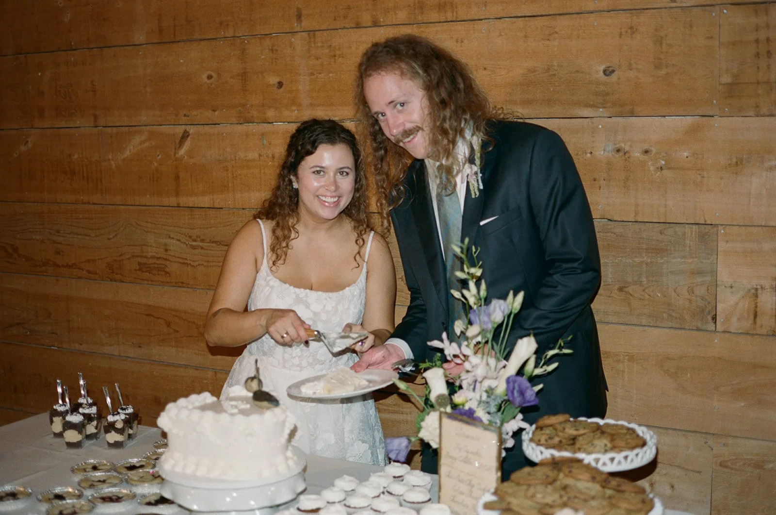 Young woman and man cutting a cake together at a celebration with desserts on the table, wooden wall background. ocracoke outerbanks wedding film photographer