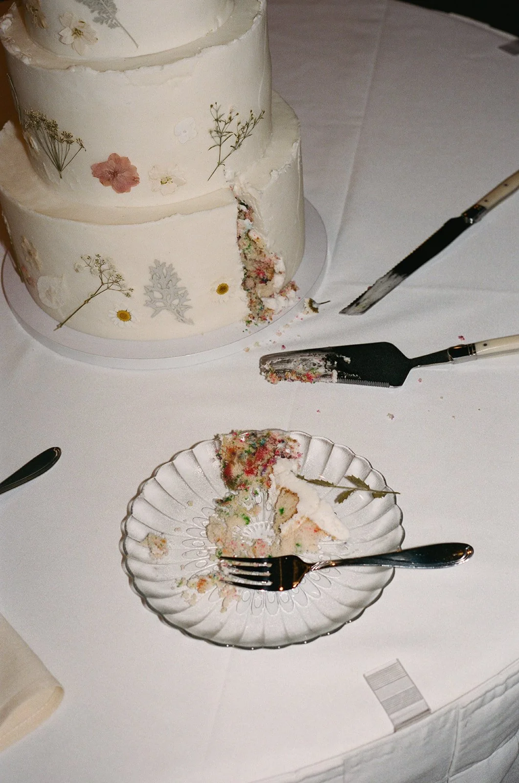 A wedding cake with floral decorations has been cut, with some slices on a glass plate, and cake serving utensils on a white tablecloth. Wintergreen resort Wedding Film Photographer