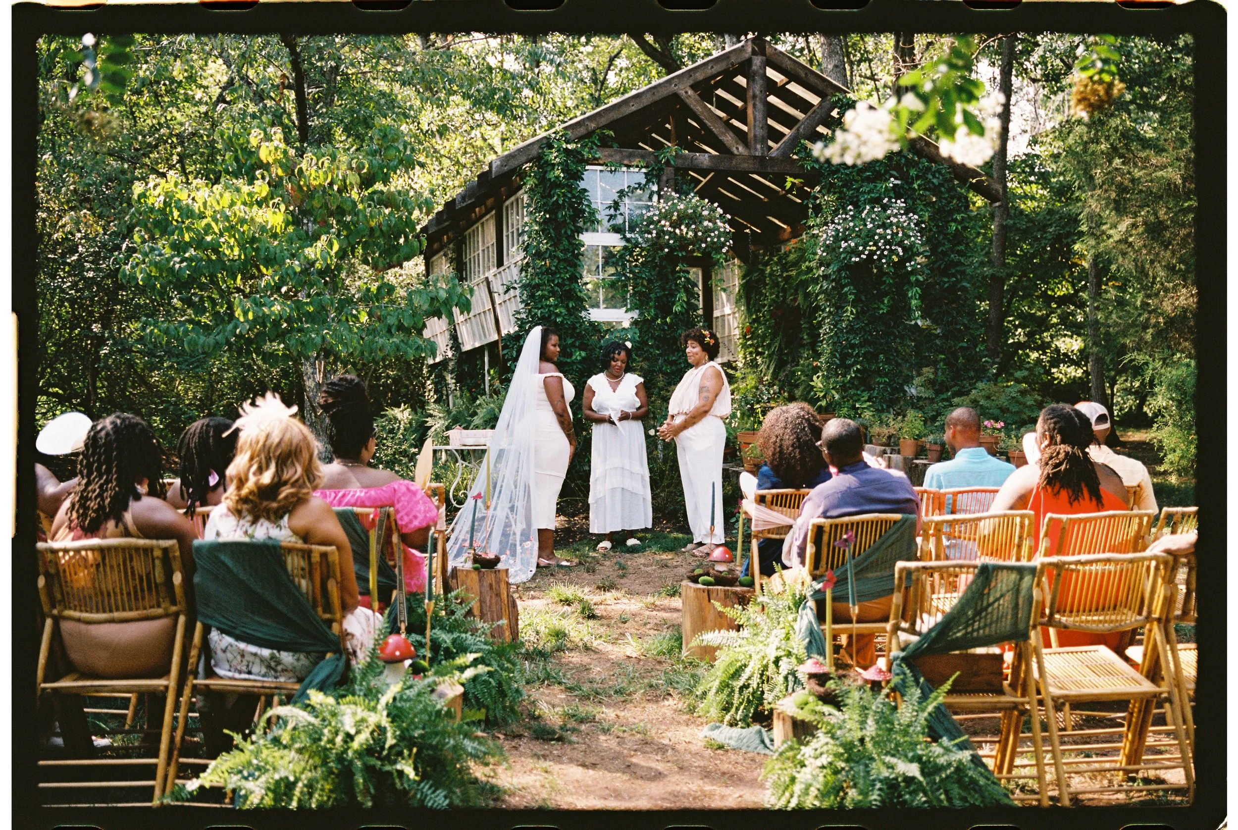 Outdoor wedding ceremony at Vaughan Greenhouse with guests seated in a garden setting, featuring a small, rustic structure adorned with greenery and flowers, and two people standing at the altar with a third officiating.